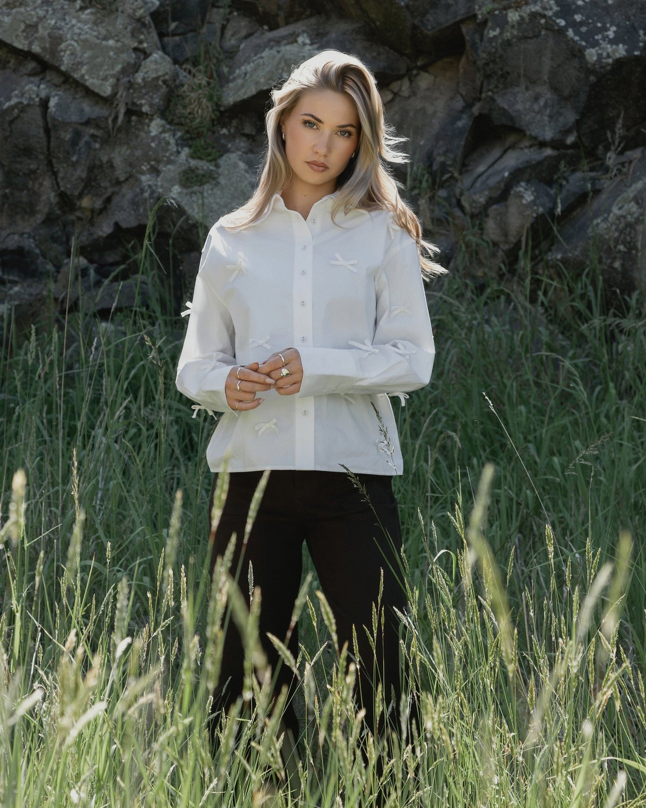 Woman in white bow-detail blouse and black pants standing in tall grass near rocks, stylish contemporary fashion