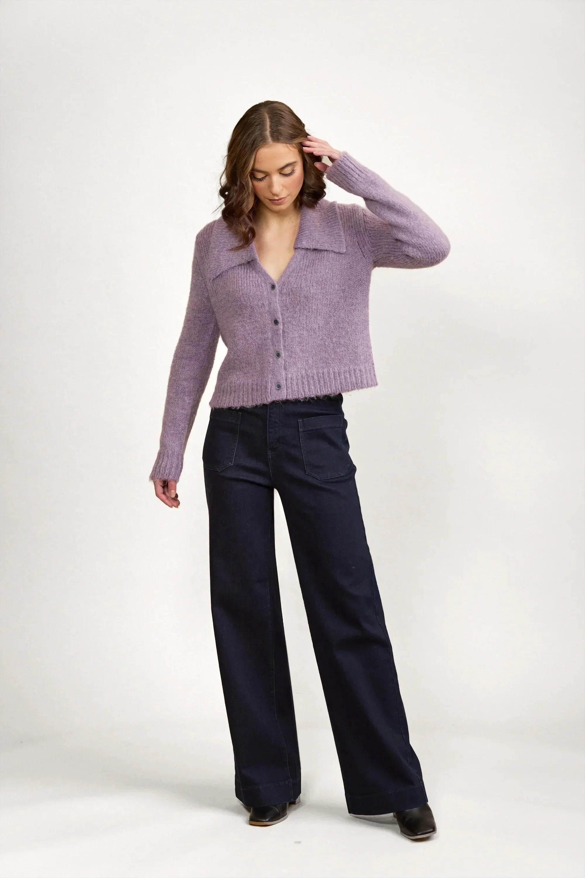 A woman with wavy brown hair wears a lavender button-up cardigan and high-waisted dark wide-leg pants, standing against a plain white background while looking down and touching her hair.