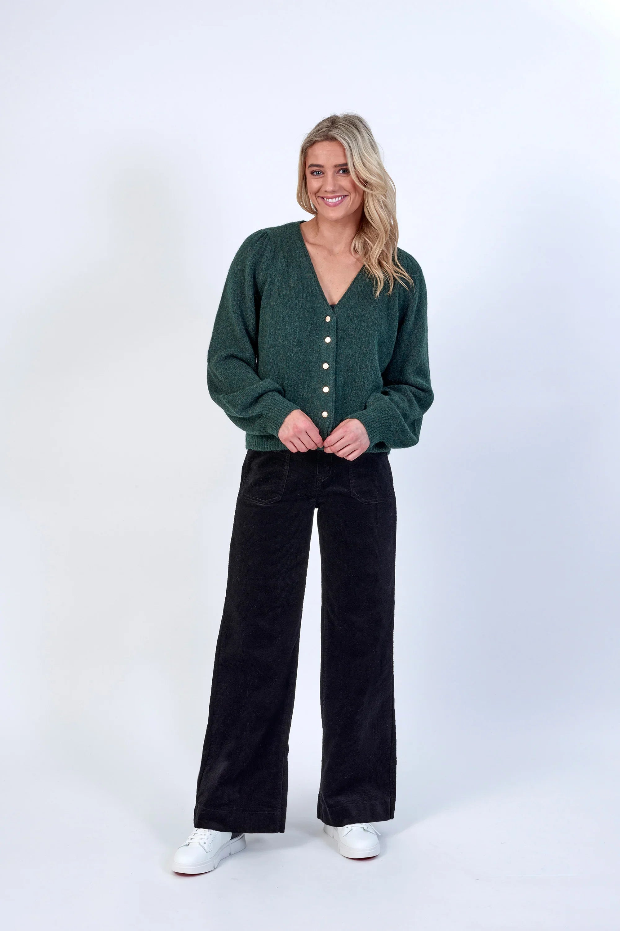 A woman with long blonde hair smiles while wearing a dark green button-up cardigan, black wide-leg pants, and white sneakers, standing against a plain white background.