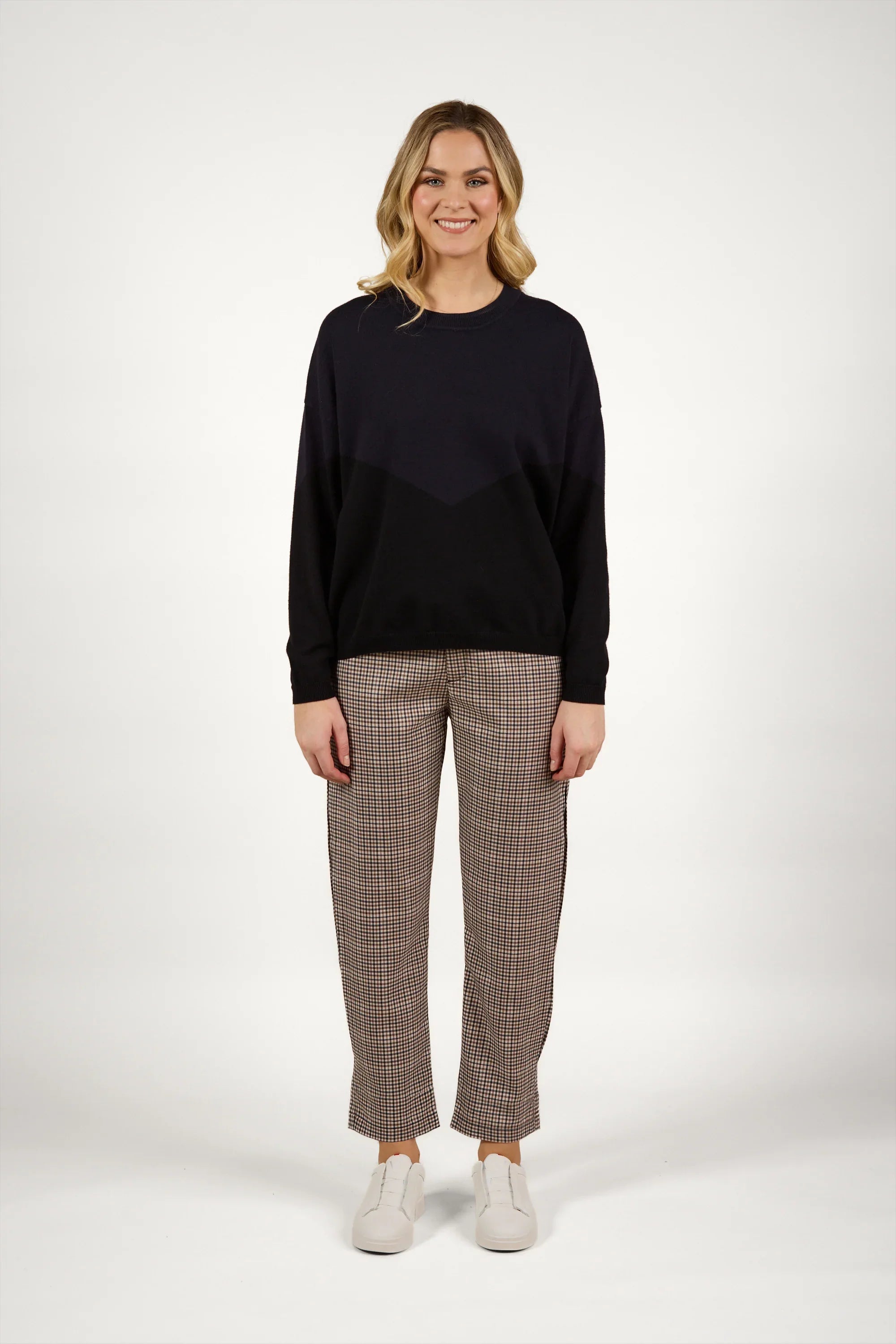 A woman stands smiling, wearing a loose black sweater, plaid trousers, and white sneakers against a plain white background.