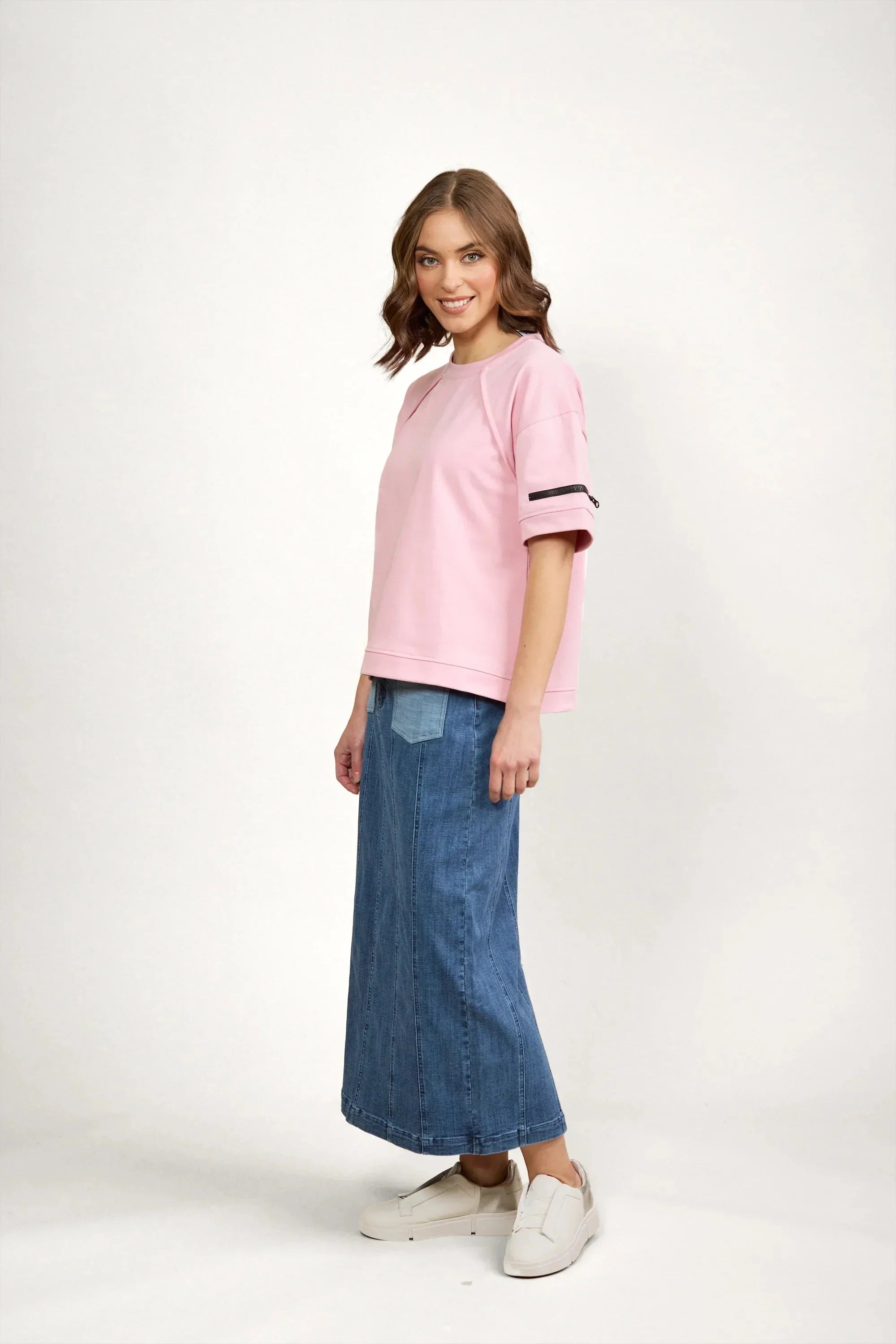 A woman stands smiling, wearing a light pink short-sleeve top with black stripes on one sleeve, a long blue denim skirt, and white slip-on shoes, against a plain white background.
