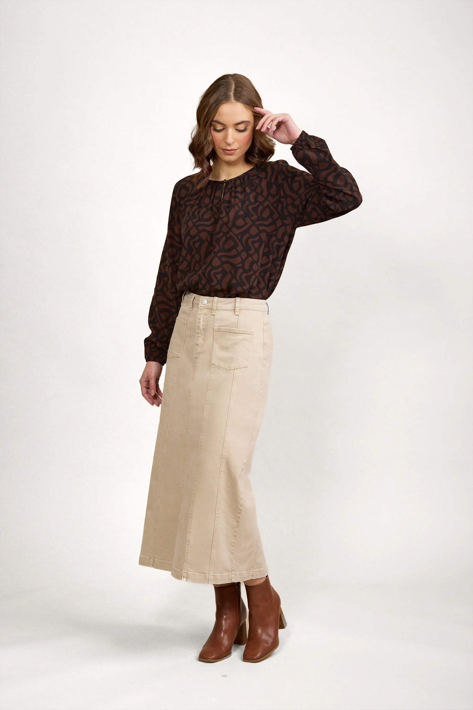 Woman standing against a white background wearing a brown patterned long-sleeve blouse, a beige midi skirt, and brown ankle boots, with one hand touching her hair.