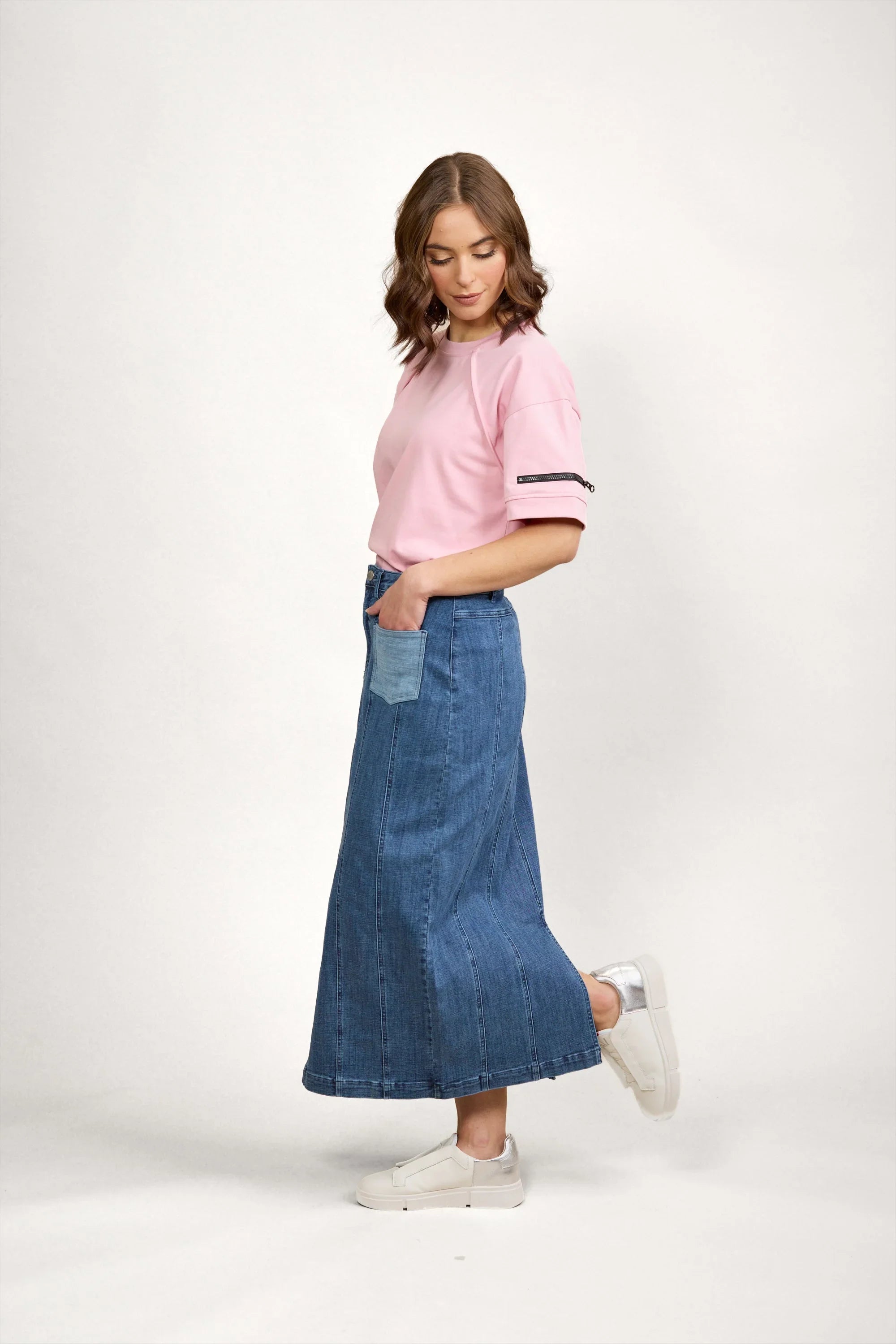 A woman stands sideways wearing a light pink short-sleeve top, a long blue denim skirt with a front pocket, and white sneakers, against a plain light background.