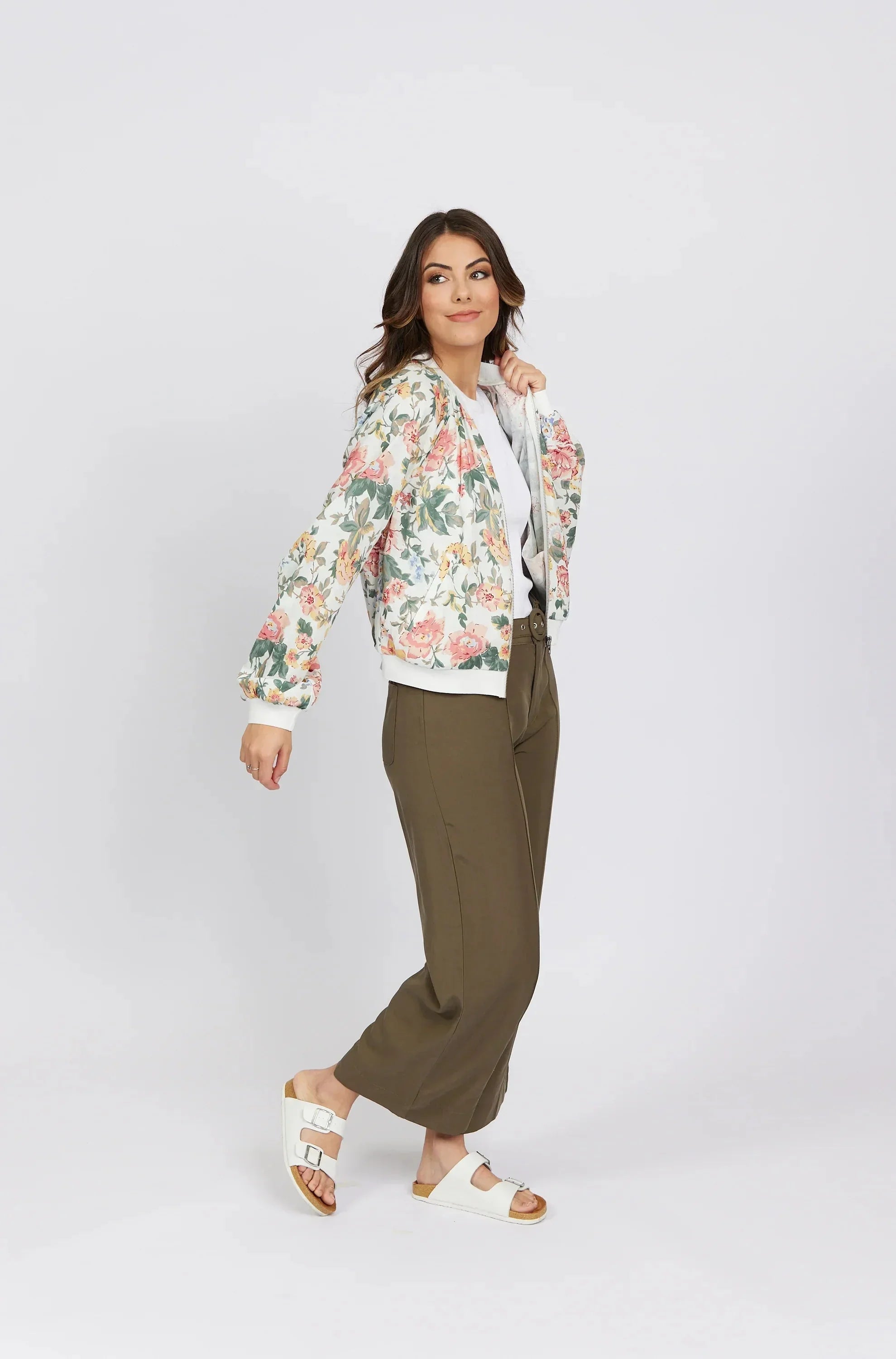 Woman posing in a floral bomber jacket, white shirt, olive green wide-leg pants, and white sandals against a plain light background. She has medium-length wavy hair and a relaxed, confident expression.