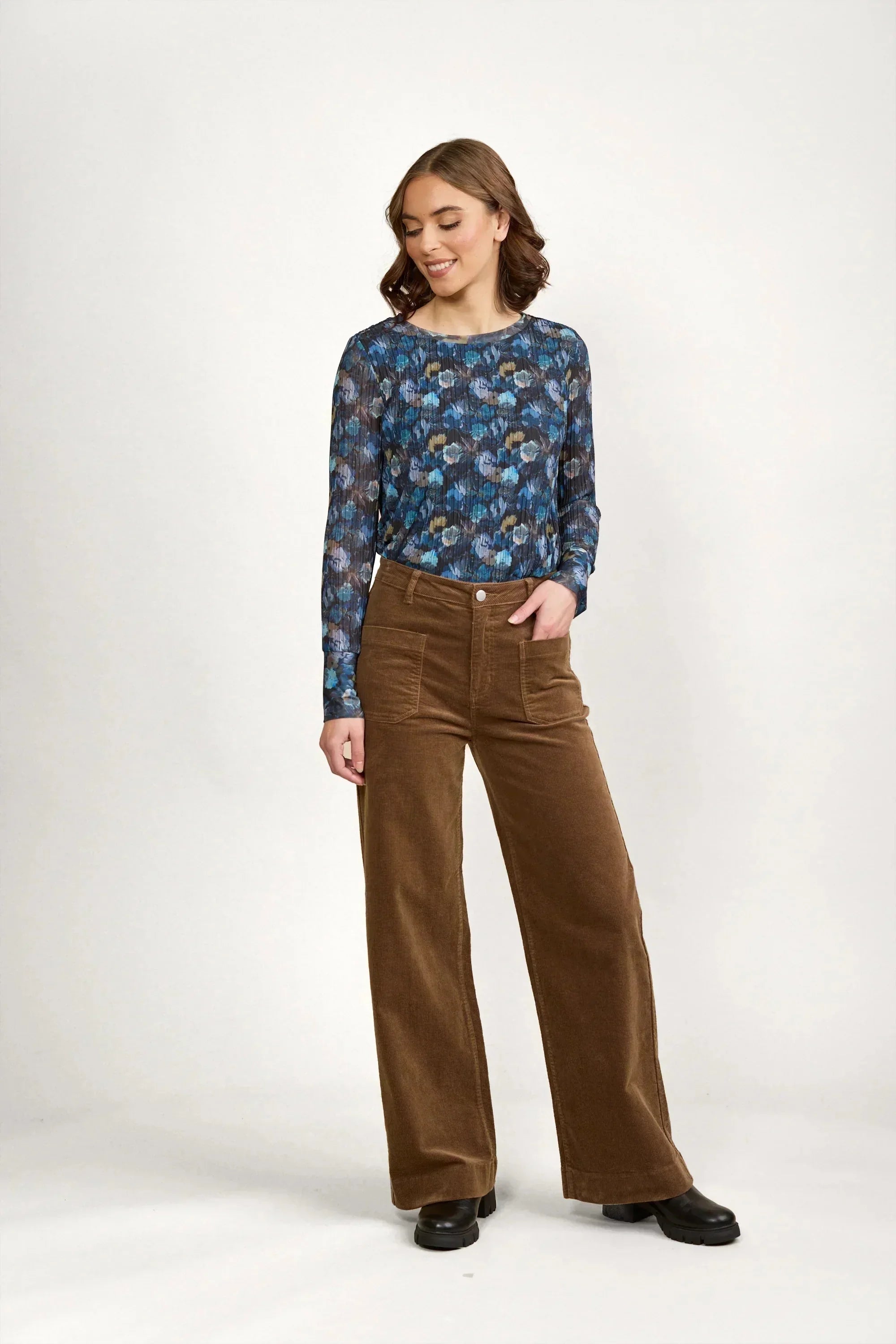 A woman stands against a plain white background, wearing a blue floral long-sleeve top and high-waisted brown wide-leg pants with black shoes. She smiles and looks to her left with one hand in her pocket.