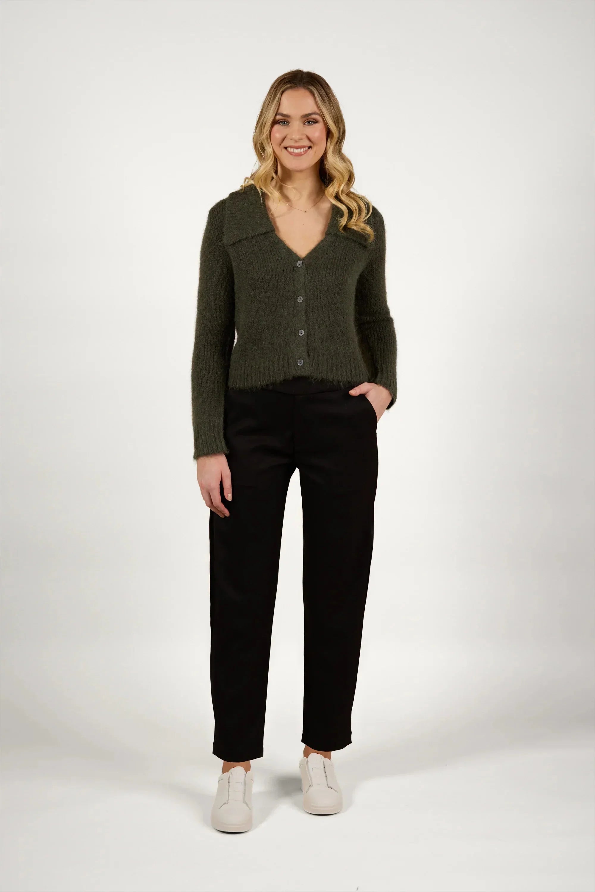 A woman with long blonde hair wears a dark green cardigan, black pants, and white sneakers. She is smiling, standing with one hand in her pocket against a plain white background.