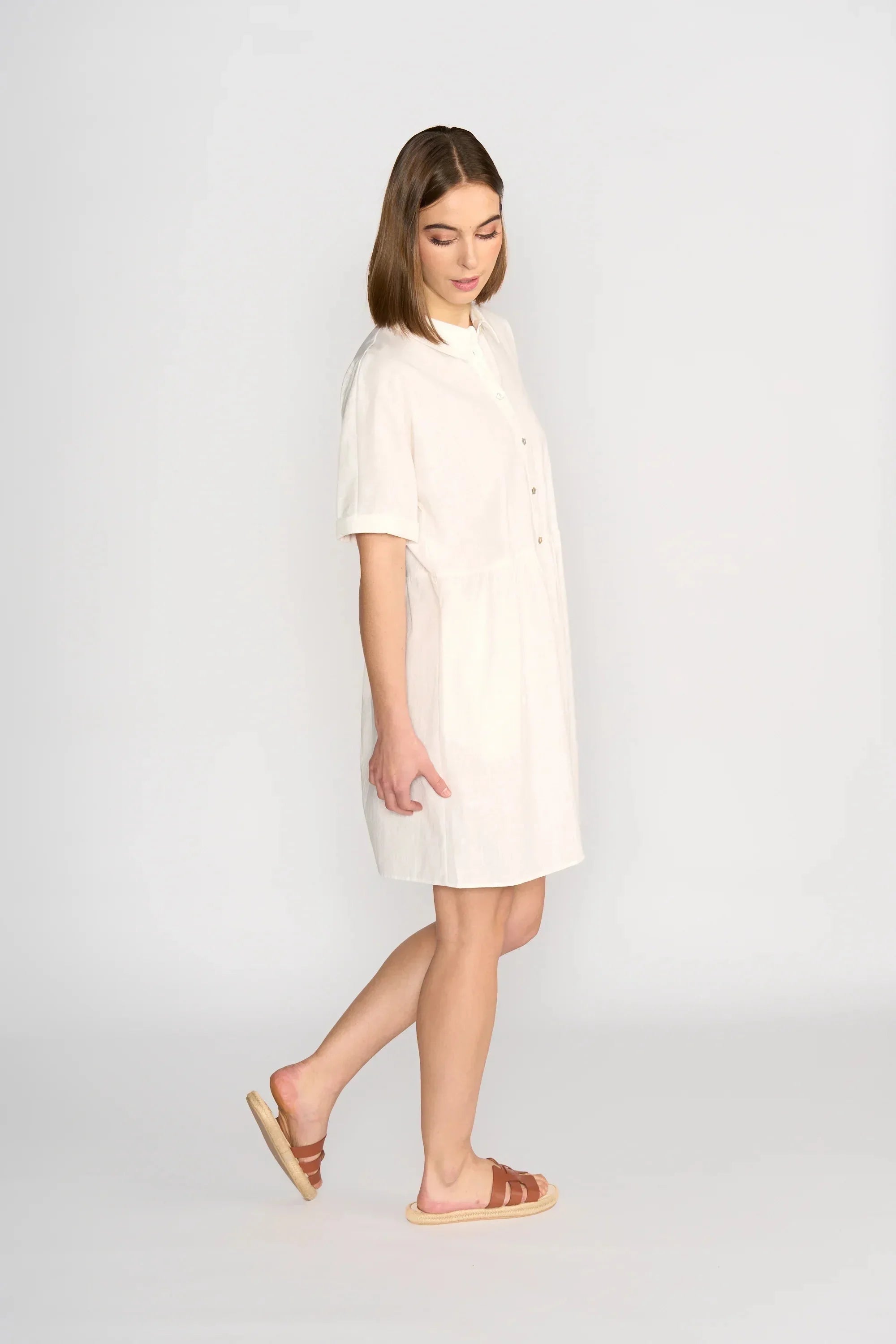 A woman wearing a loose, short-sleeved, knee-length white shirt dress and brown flat sandals stands against a plain light background, looking down and slightly to the side.