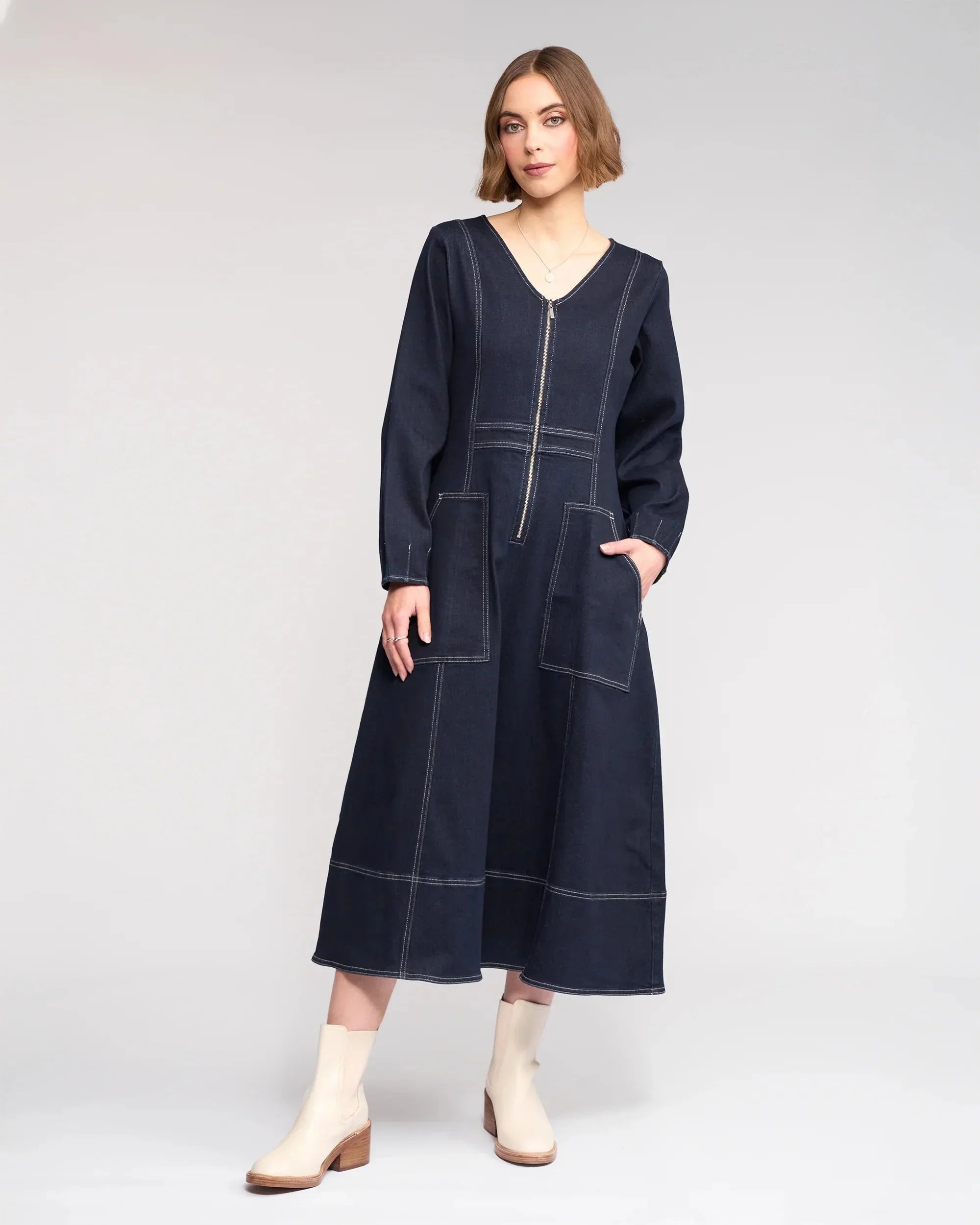 A woman wearing a long-sleeve, dark denim midi dress with white contrast stitching, front zipper, and large front pockets stands confidently in light ankle boots against a plain background.