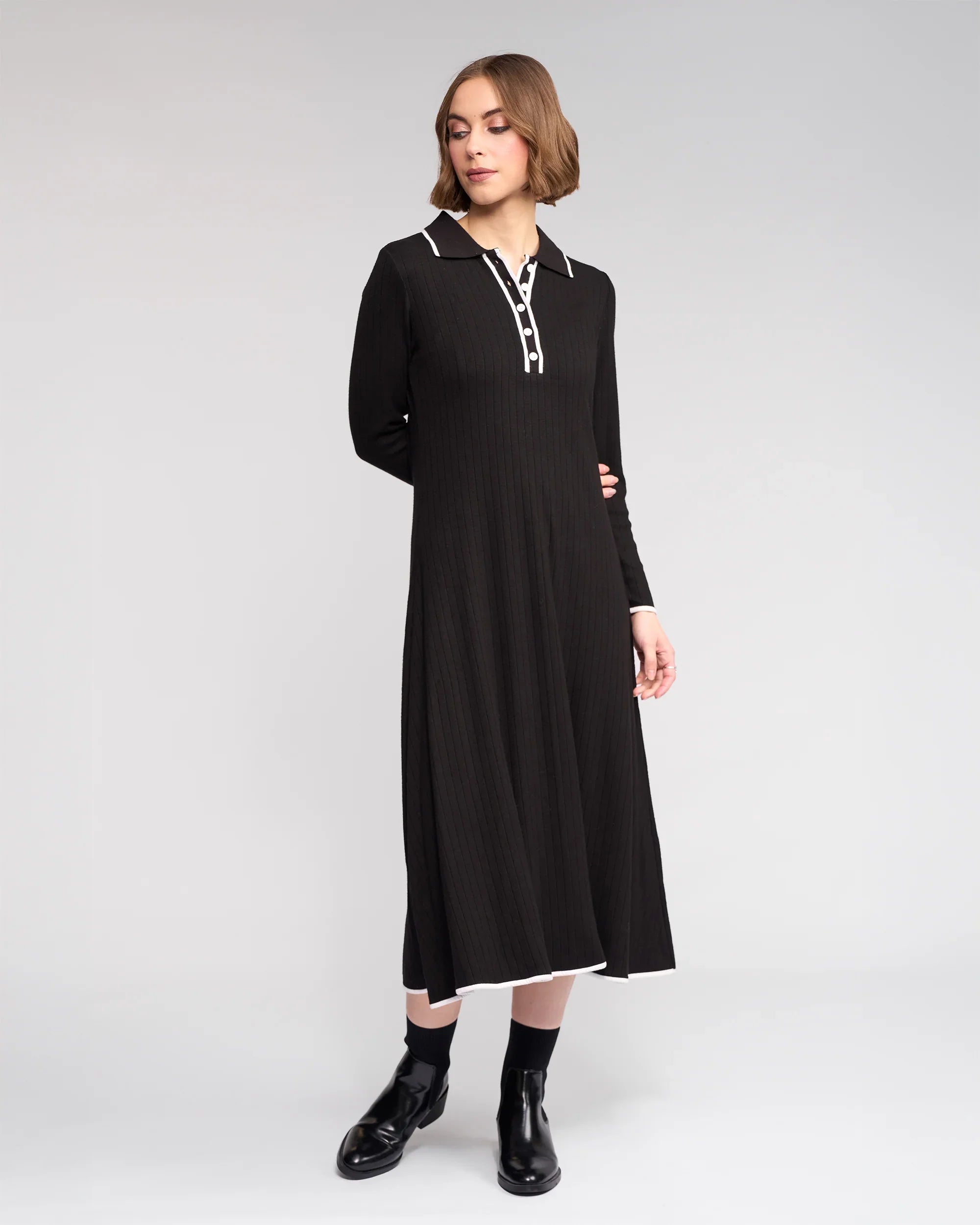 A woman stands against a light gray background wearing the Knewe Label Ritual Dress in black—a ribbed knit piece with a shirt-style collar, white trim, and black ankle boots for a chic winter look.