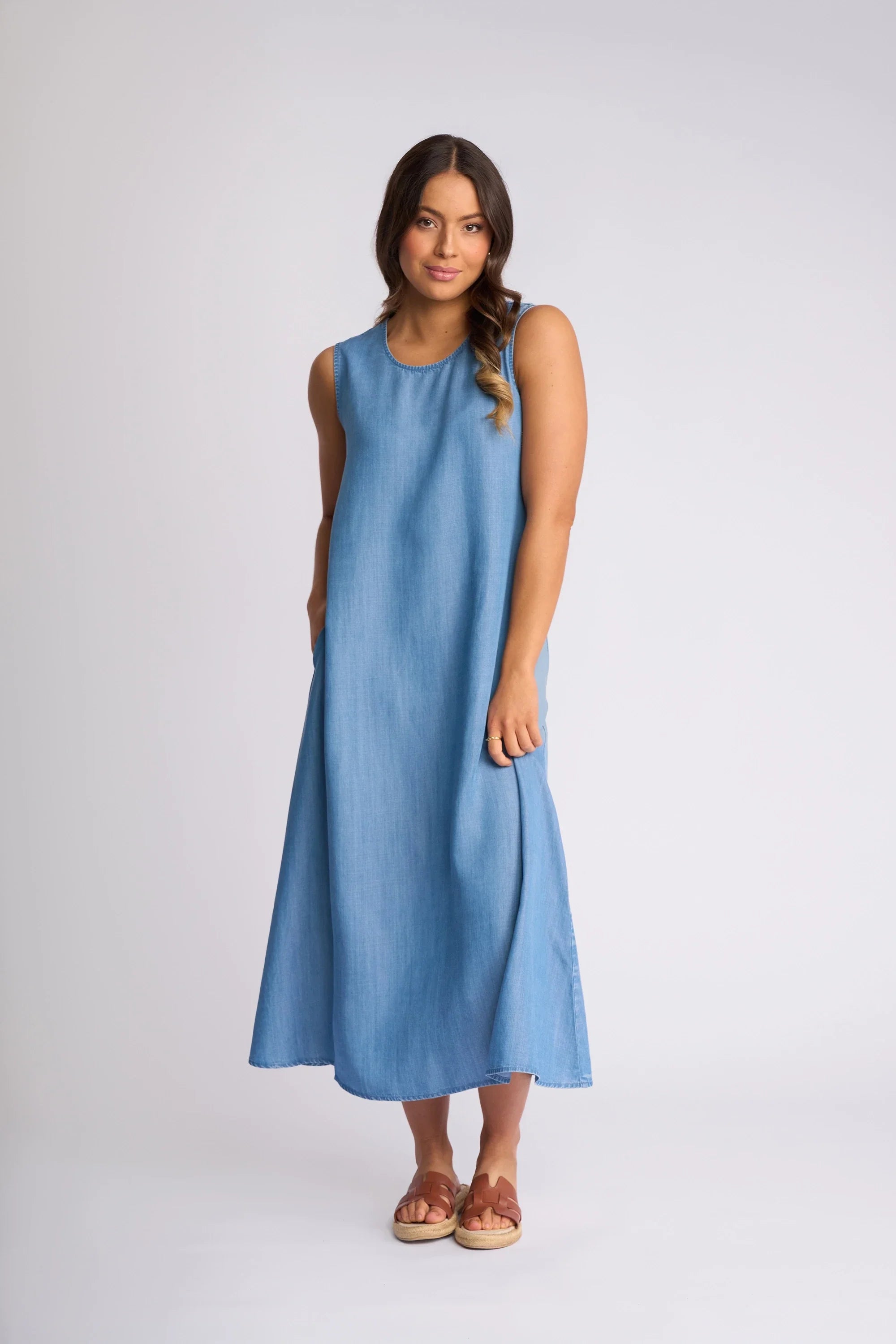 A woman wearing a long-sleeve, dark denim midi dress with white contrast stitching, front zipper, and large front pockets stands confidently in light ankle boots against a plain background.