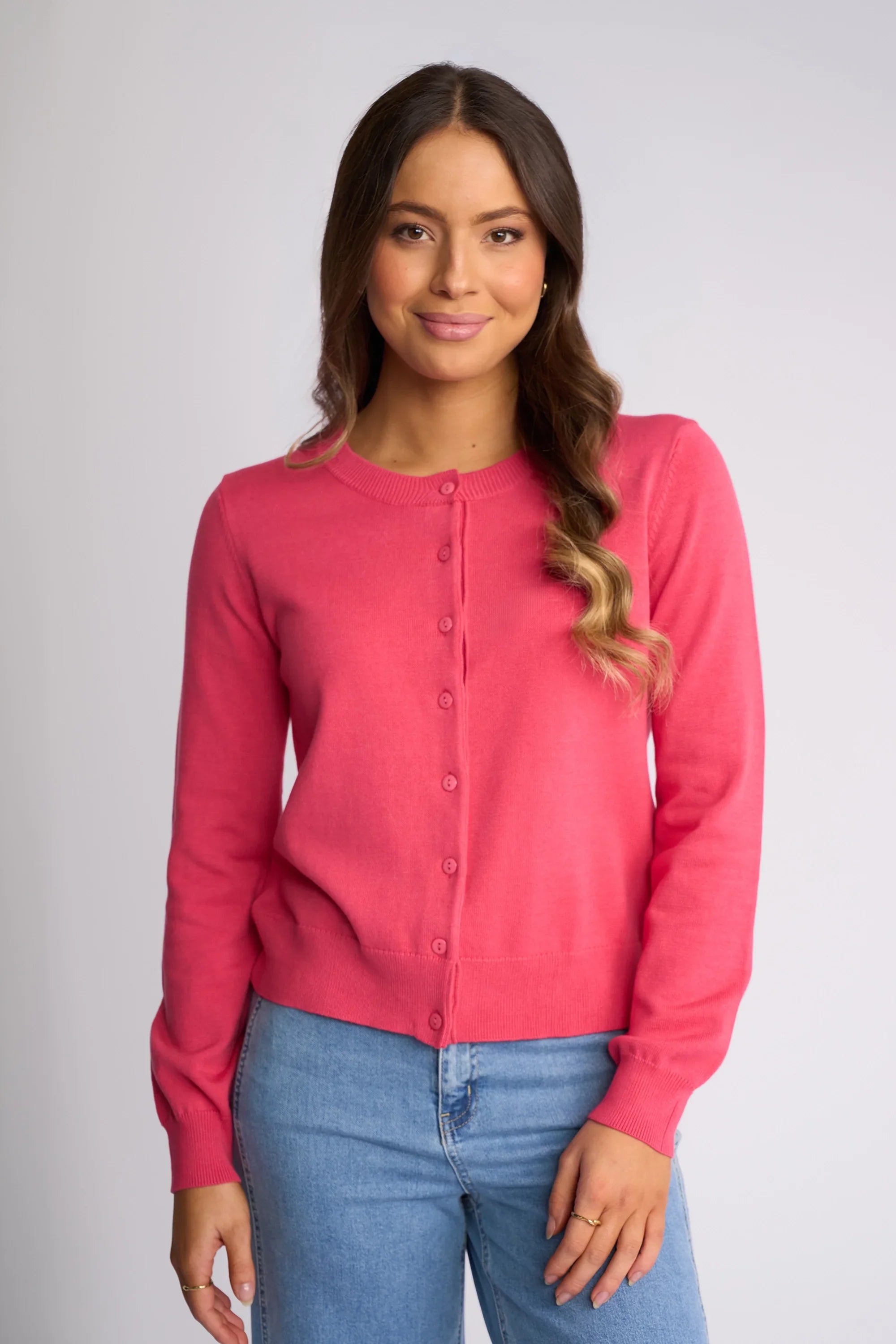 A woman with wavy brown hair wears a lavender button-up cardigan and high-waisted dark wide-leg pants, standing against a plain white background while looking down and touching her hair.
