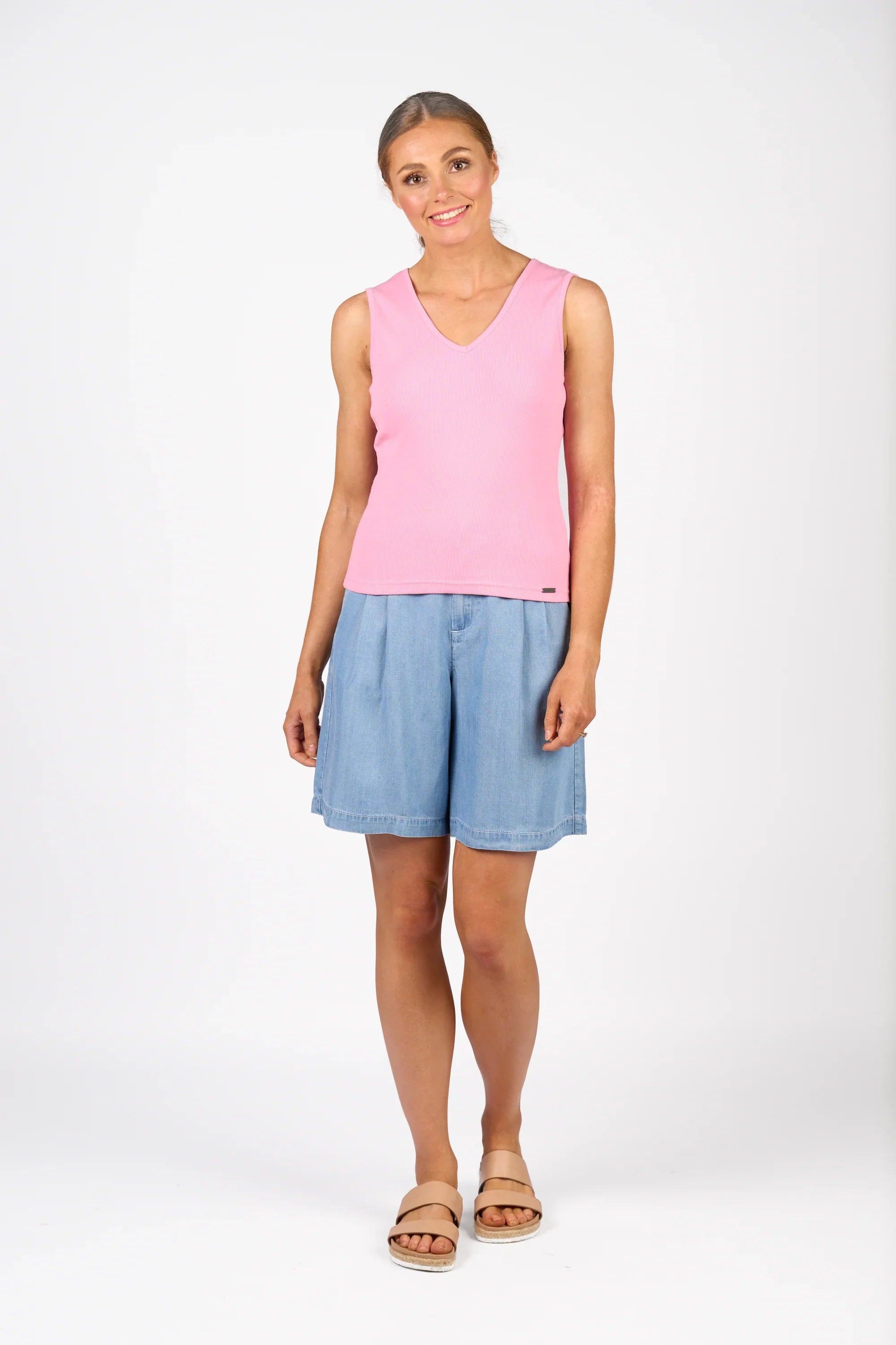 A woman stands smiling, wearing a sleeveless pink top, light blue denim shorts, and tan sandals against a plain white background.