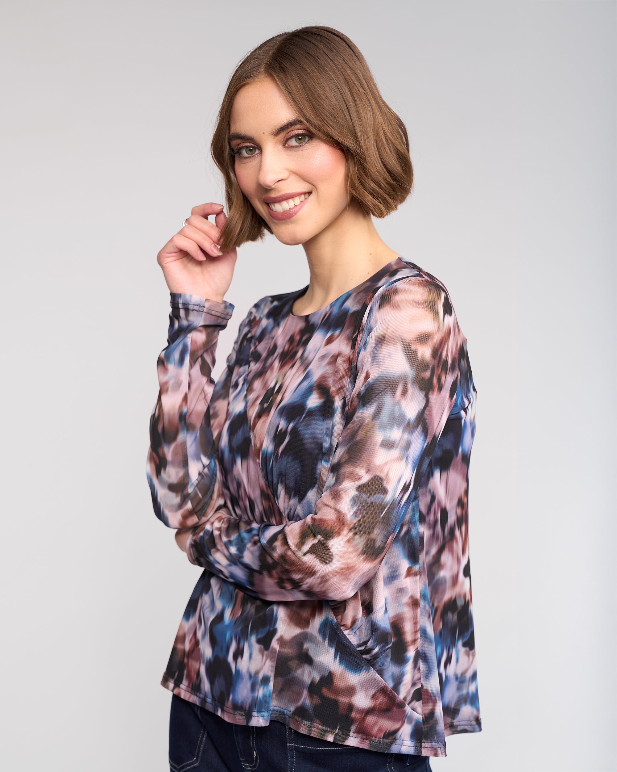 A woman with short brown hair smiles with one hand near her face, wearing the Apex Top - Celine by Knewe Label—an abstract print mesh long sleeve—paired with dark jeans, against a light gray background.