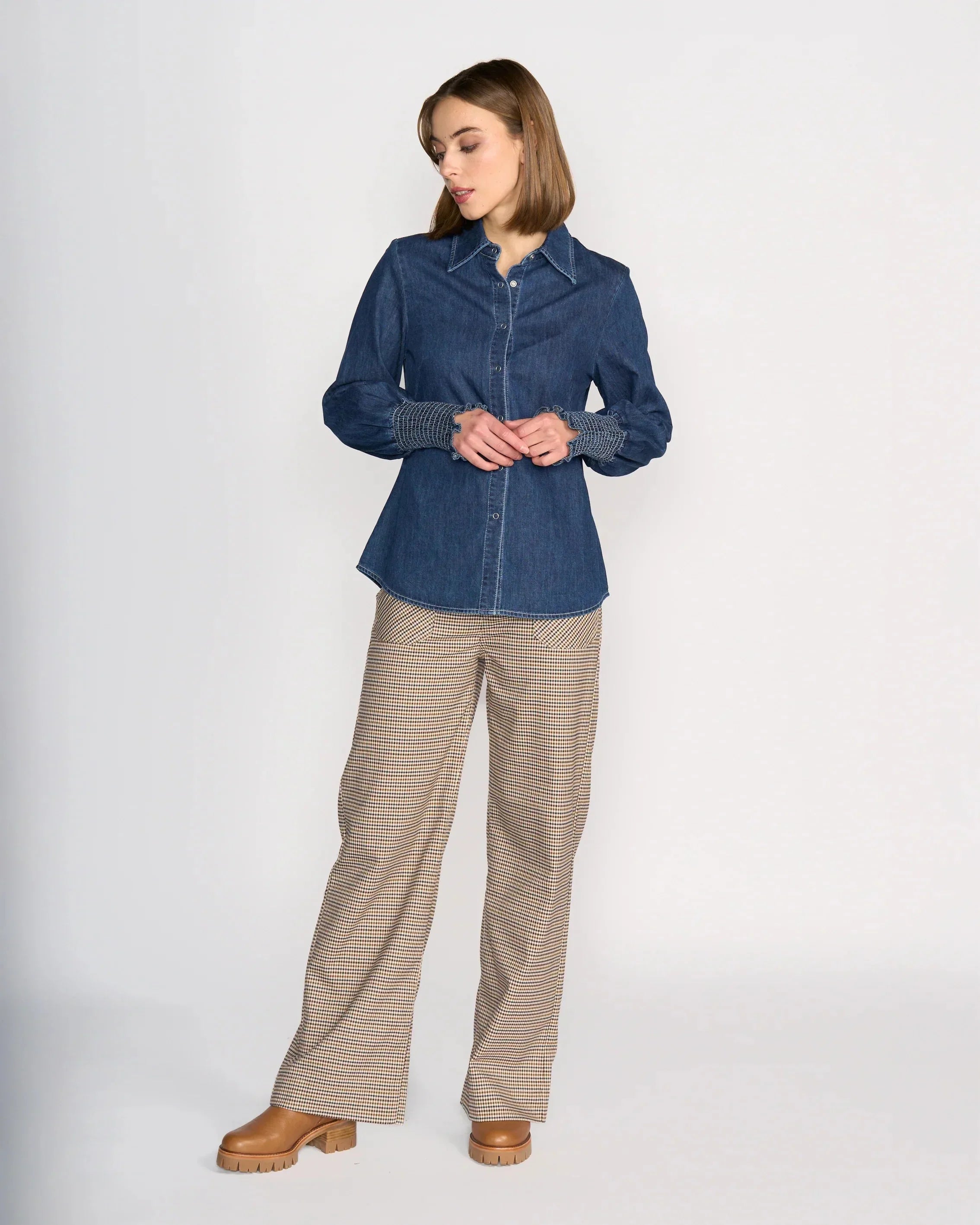 A woman stands against a white background wearing a blue denim button-up shirt, beige plaid wide-leg pants, and tan platform shoes, looking down and touching her wrist with one hand.