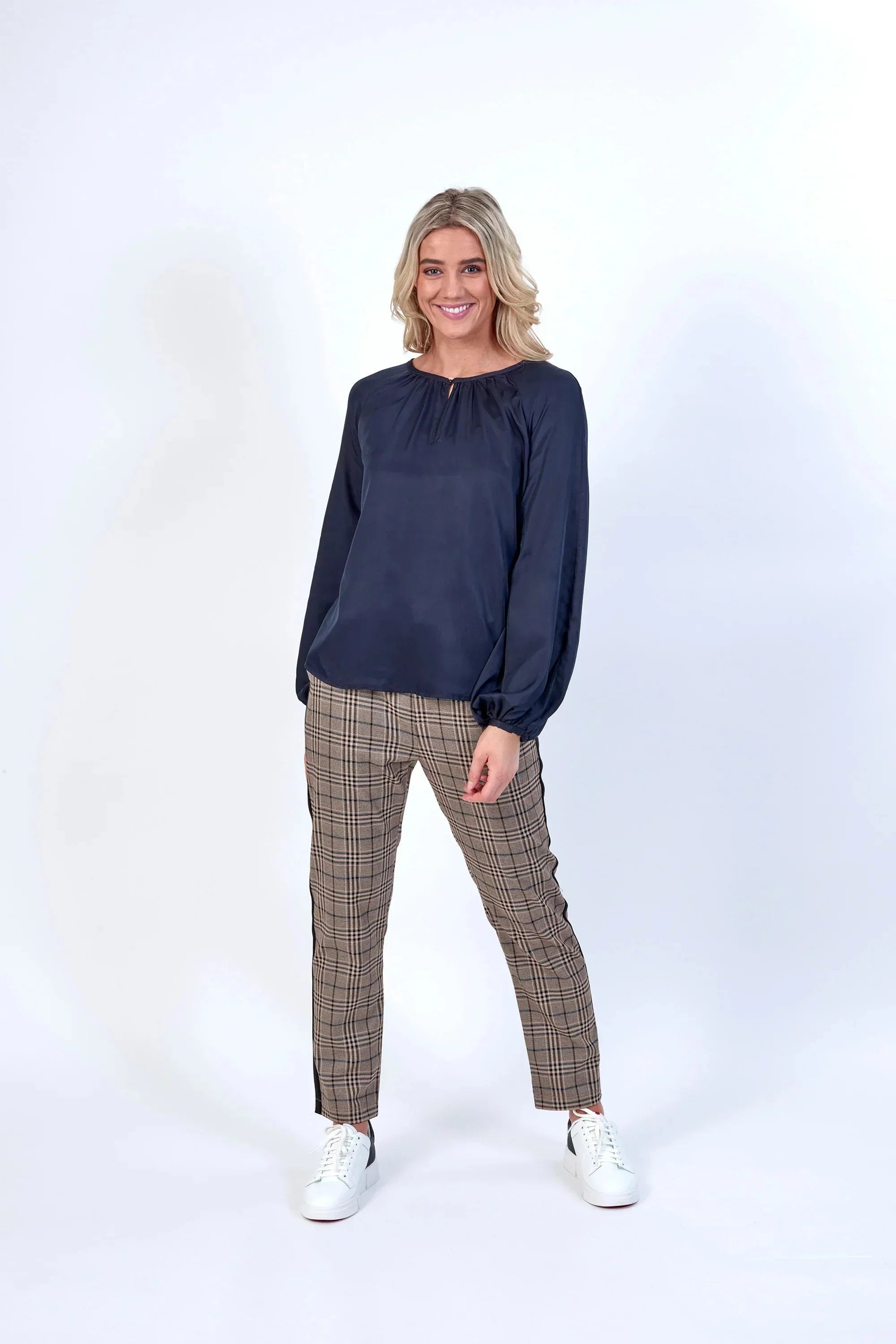 A woman with blonde hair is standing and smiling, wearing a navy long-sleeve blouse, plaid pants, and white sneakers against a plain white background.