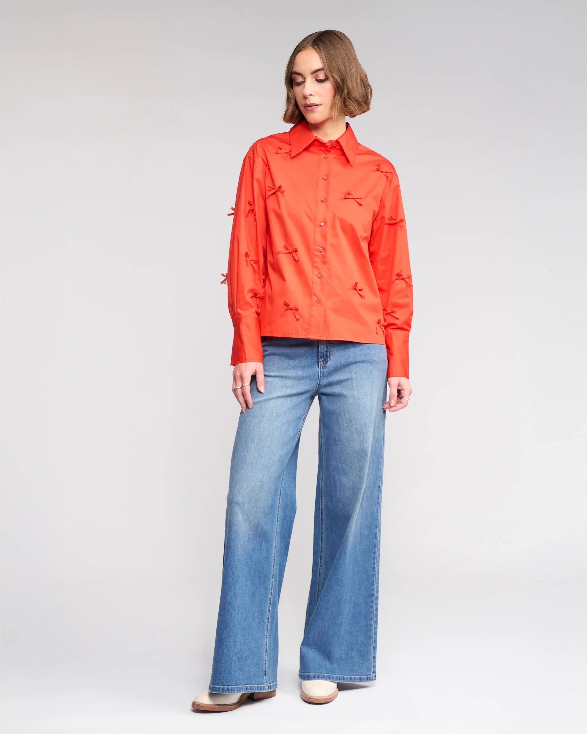 A person wearing a bright orange long-sleeve shirt with textured details and wide-leg blue jeans stands against a plain light background, looking slightly downward.