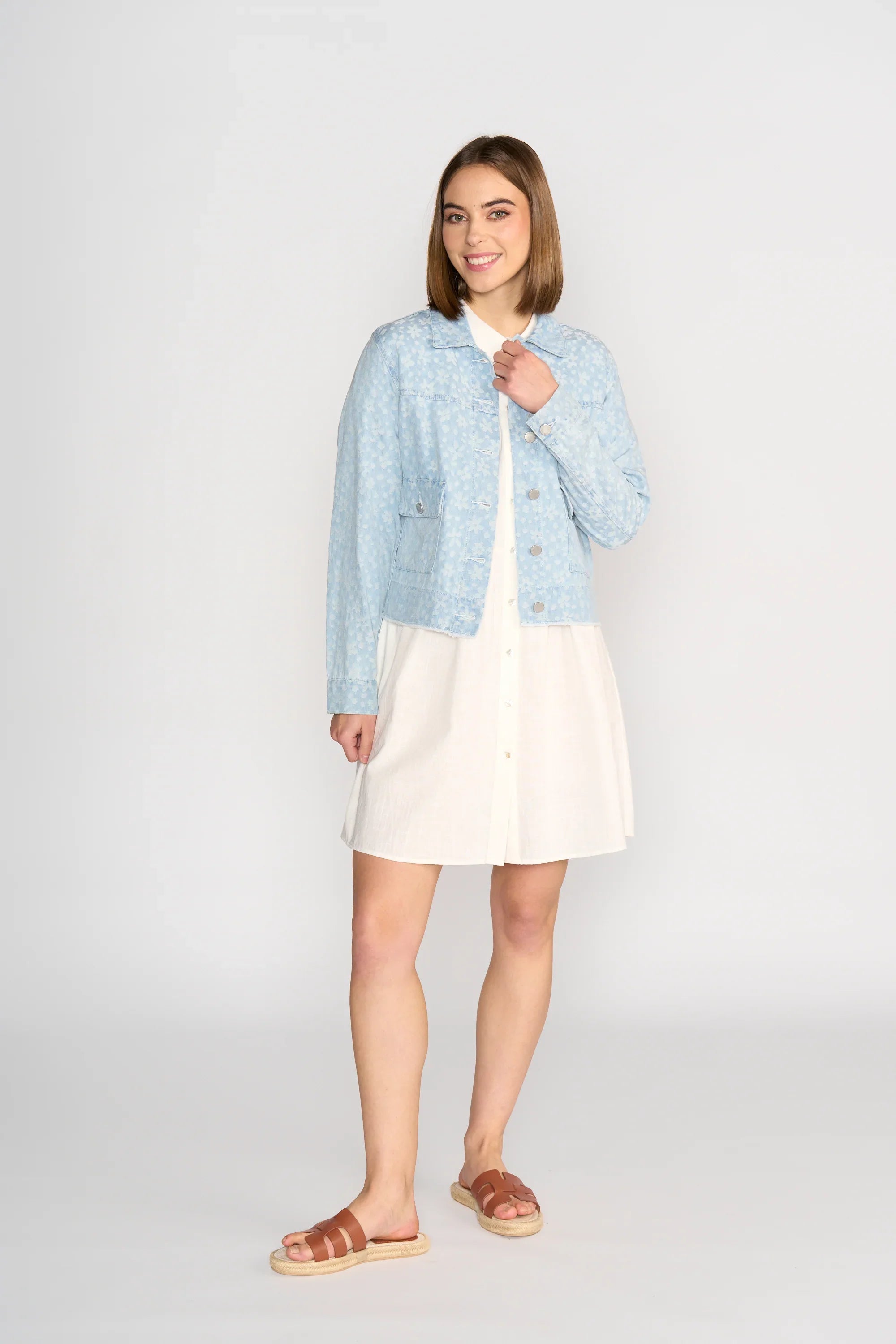 A woman stands smiling, wearing a light blue patterned jacket over a white button-down dress and brown sandals, posing in front of a plain light background.