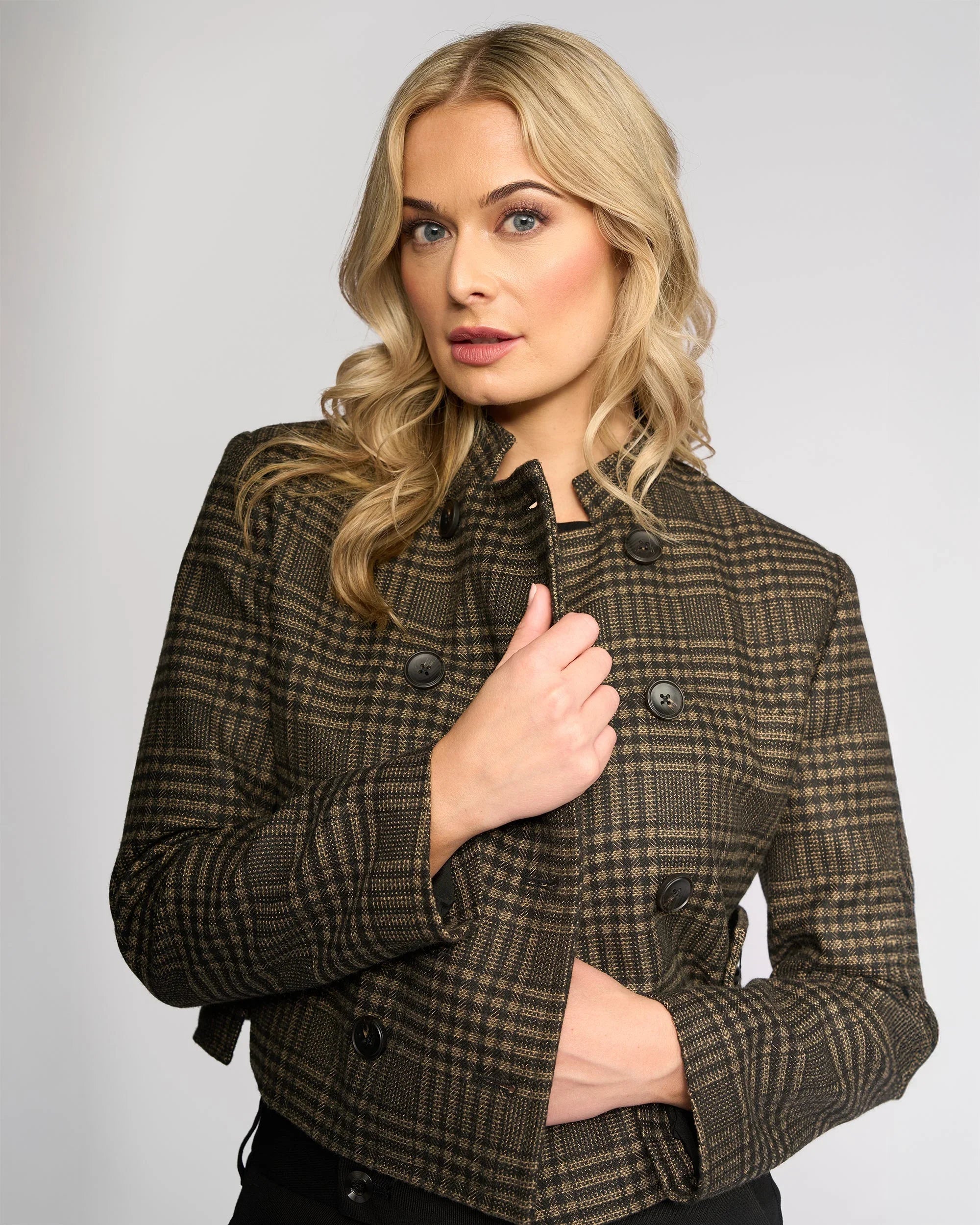 A woman with long blonde hair, wearing a brown and black plaid buttoned jacket, poses confidently with one hand on her jacket against a plain light gray background.