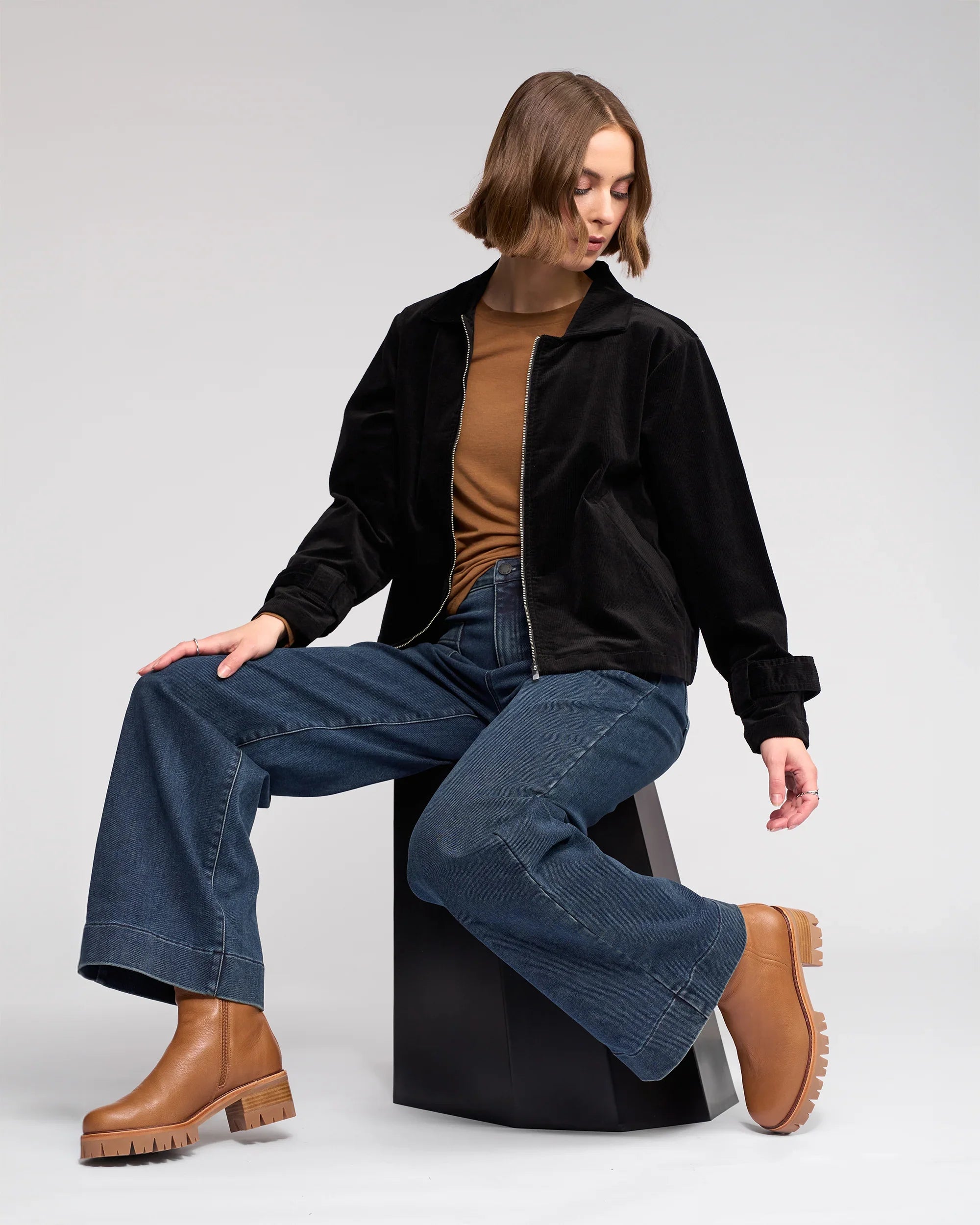 A person with a bob haircut wears a brown shirt, black jacket, wide-leg blue jeans, and tan platform boots while sitting on a black geometric stool against a plain background.
