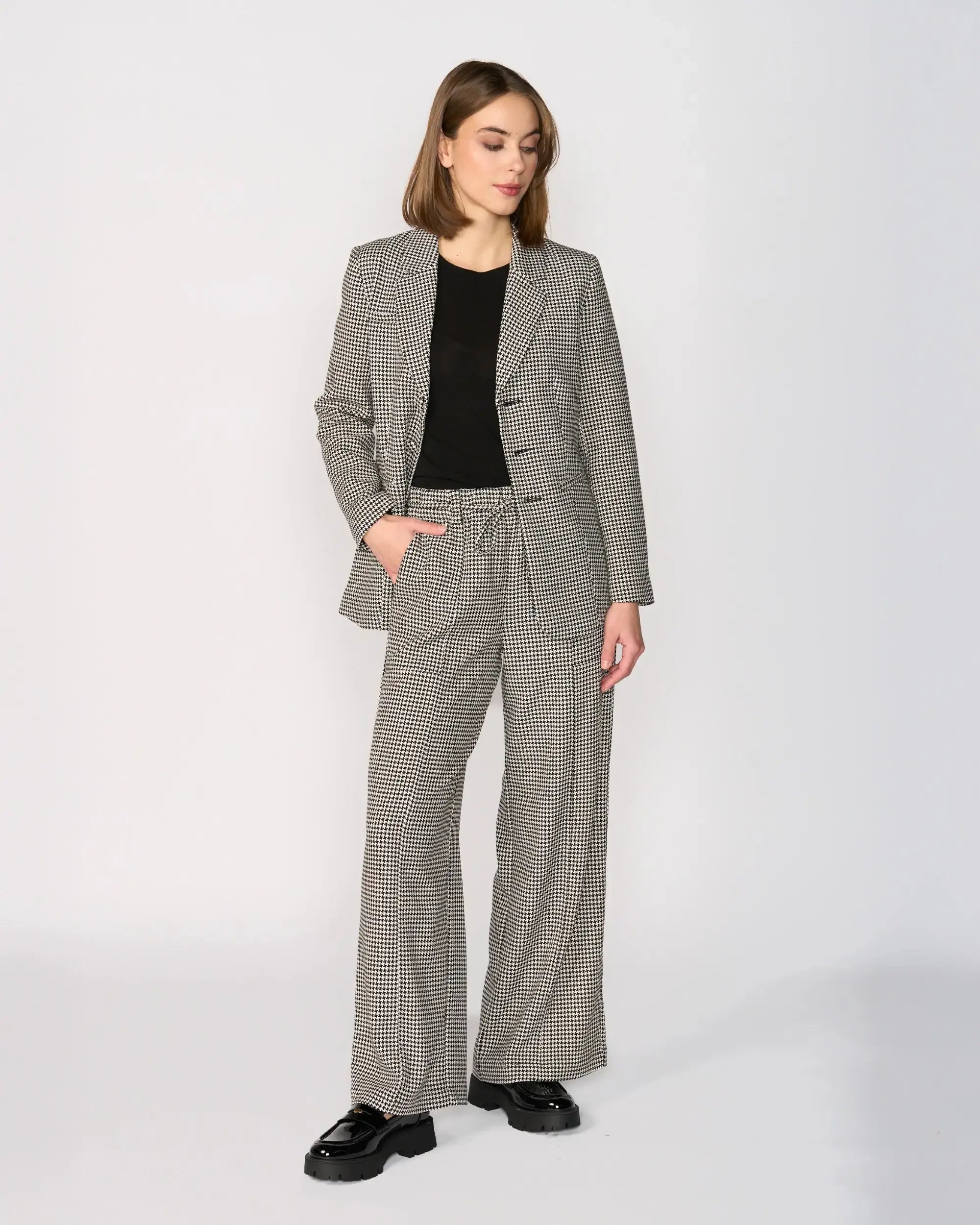 A woman stands smiling, wearing a dark long-sleeve sweater with a white stripe on the arm, plaid wide-leg trousers, and black boots, against a plain white background.