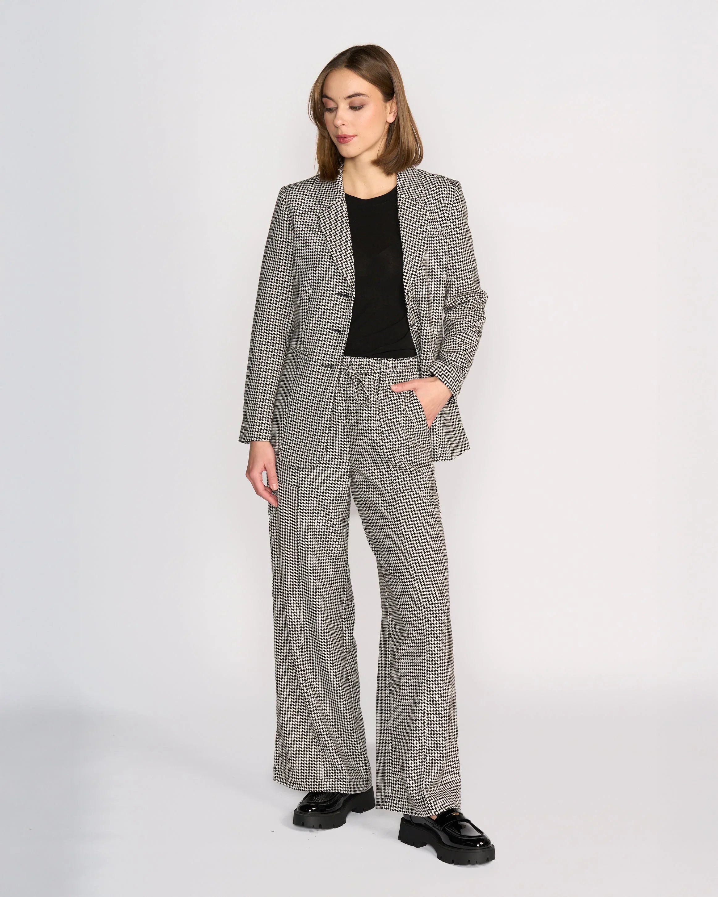 Woman standing against a plain background, wearing a black top, black-and-white checkered blazer and matching wide-leg trousers, with one hand in her pocket and black shoes.