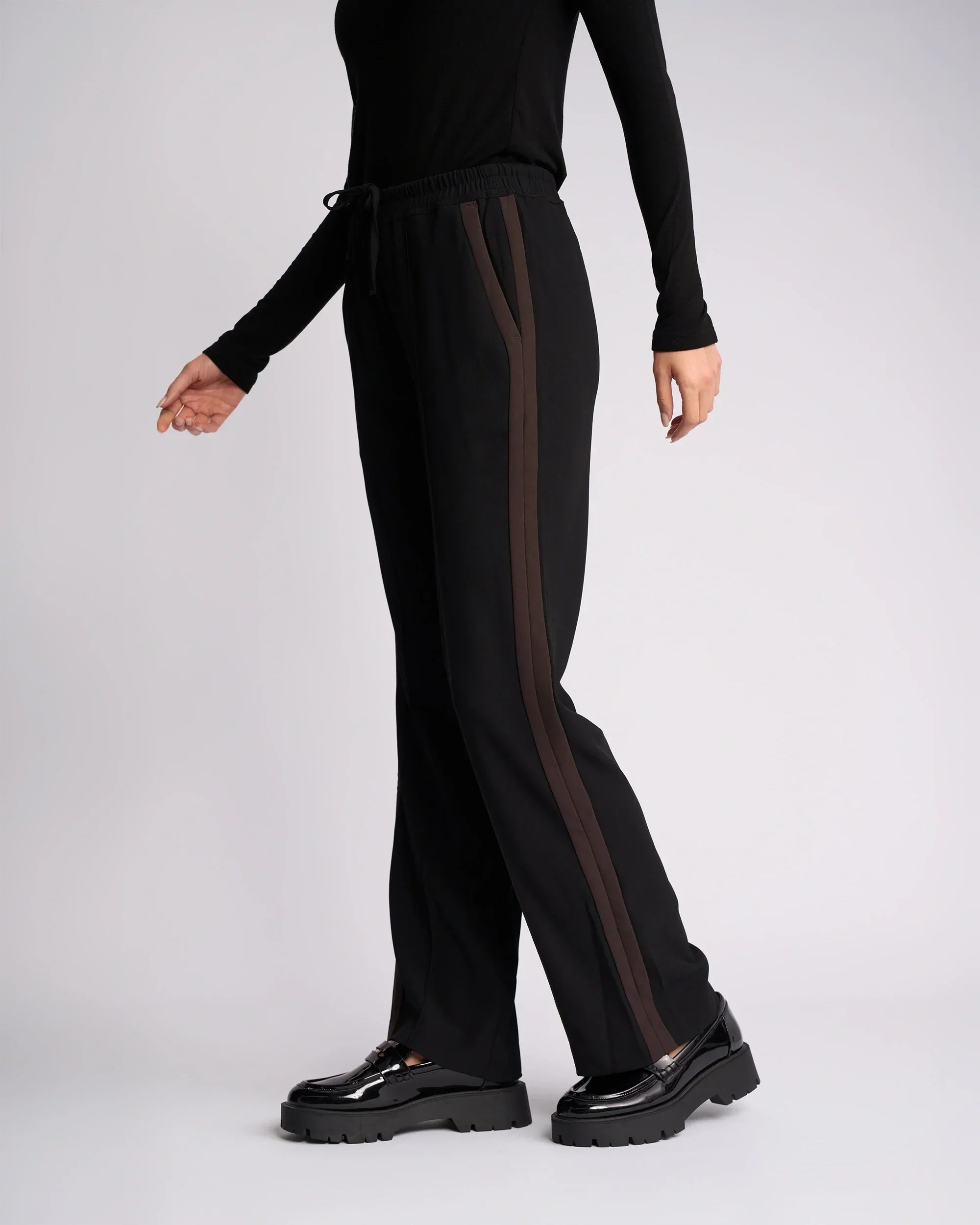 A person wearing black drawstring pants with brown side stripes, a black long-sleeve top, and black platform shoes stands against a light gray background.