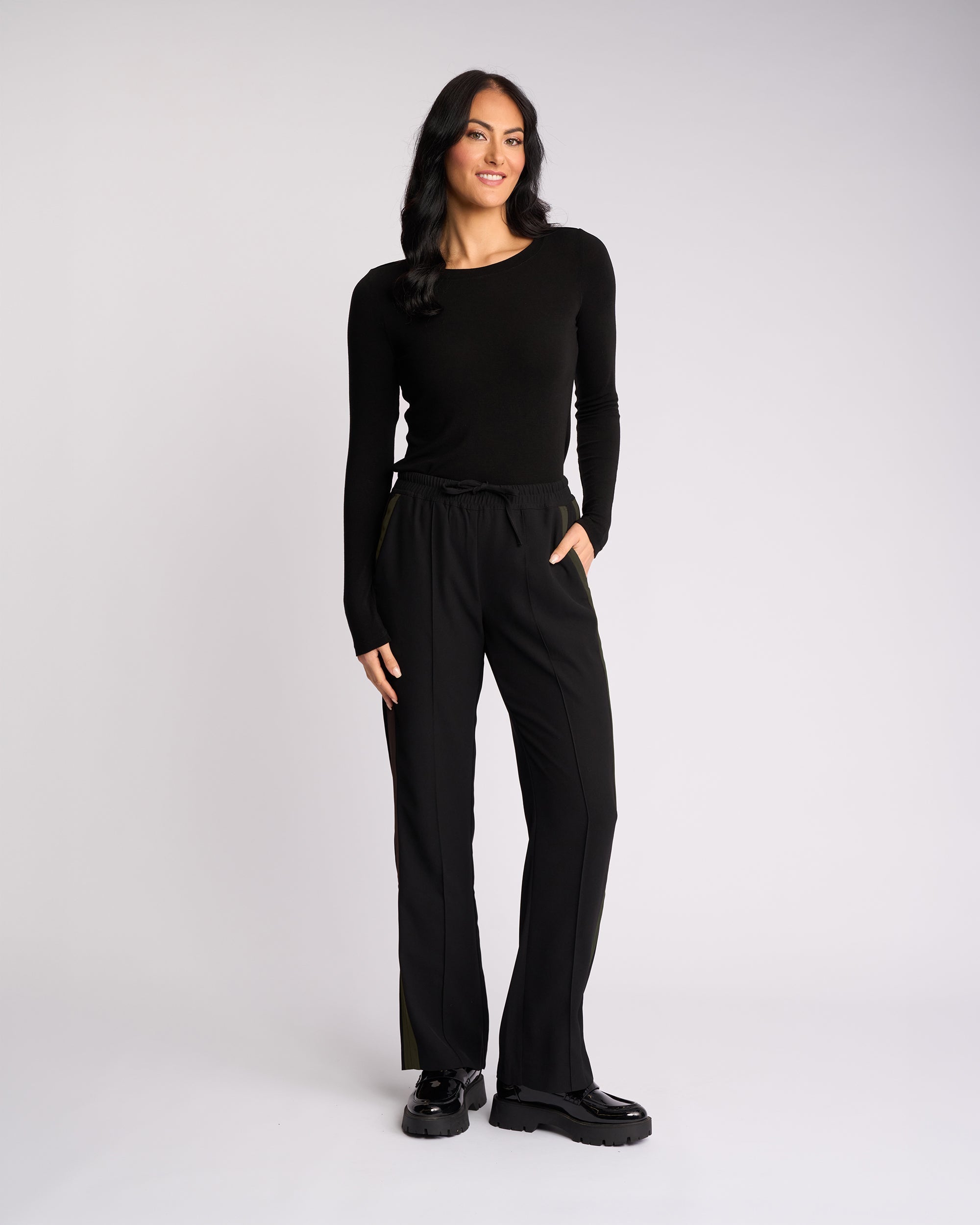 A woman with long dark hair smiles against a plain light background, wearing the Knewe Label Bowie Pant in Black/Moss with an elasticated waistband, a black long-sleeve top, black shoes, and one hand in her pocket.