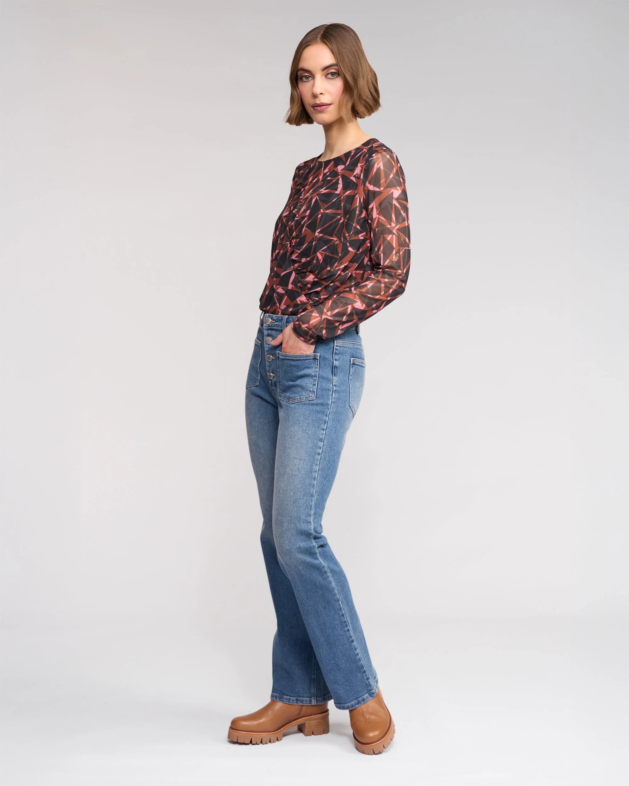 A woman stands sideways, wearing a geometric-patterned long-sleeve top, blue jeans, and brown platform shoes, against a plain light gray background.