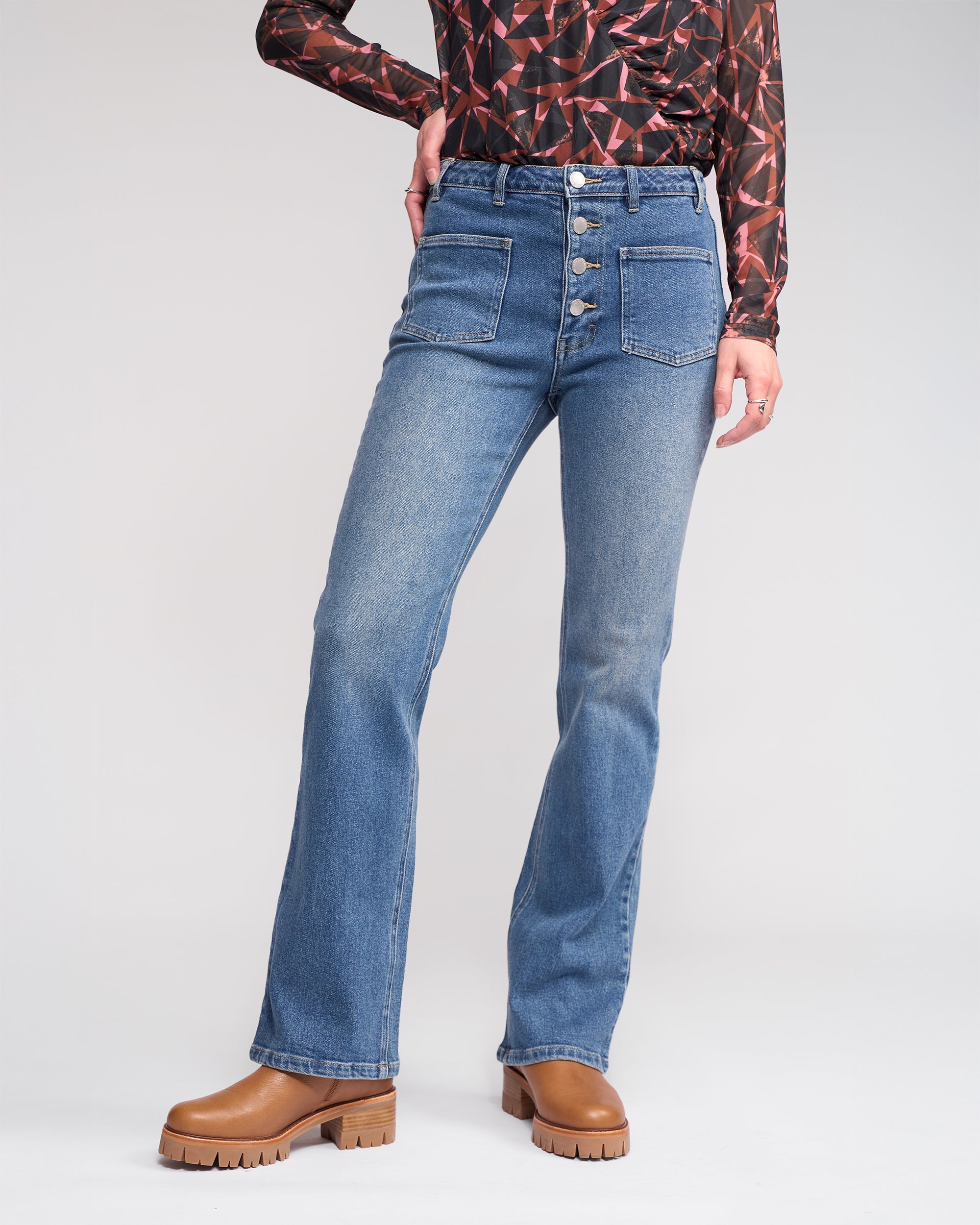 A person stands against a plain background wearing Knewe Label's Potter Jean - Vintage Denim, styled with a patterned long-sleeve top and brown platform shoes.