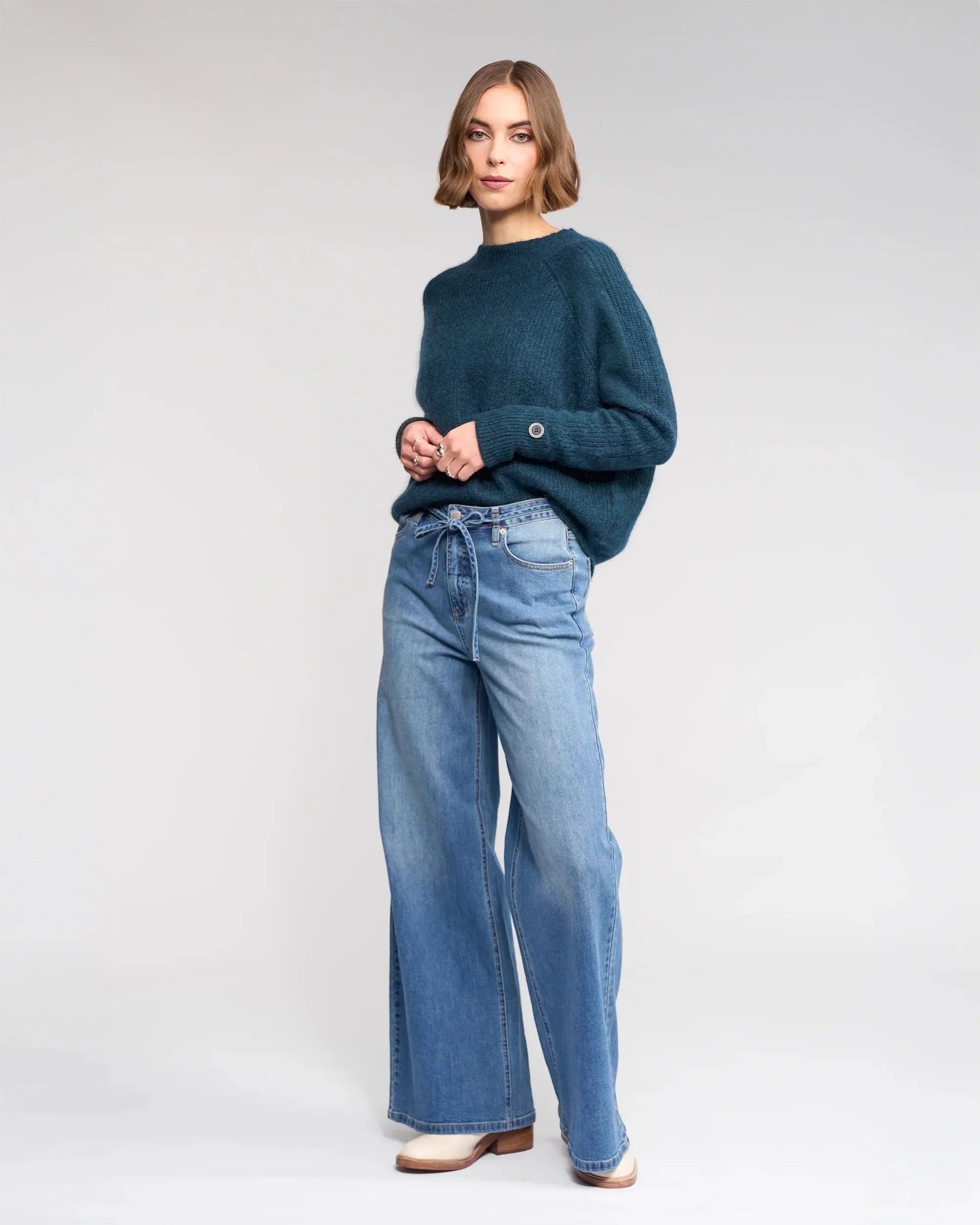 Woman standing against a plain background, wearing a teal knit sweater and high-waisted, wide-leg blue jeans with a tie belt, paired with beige shoes.