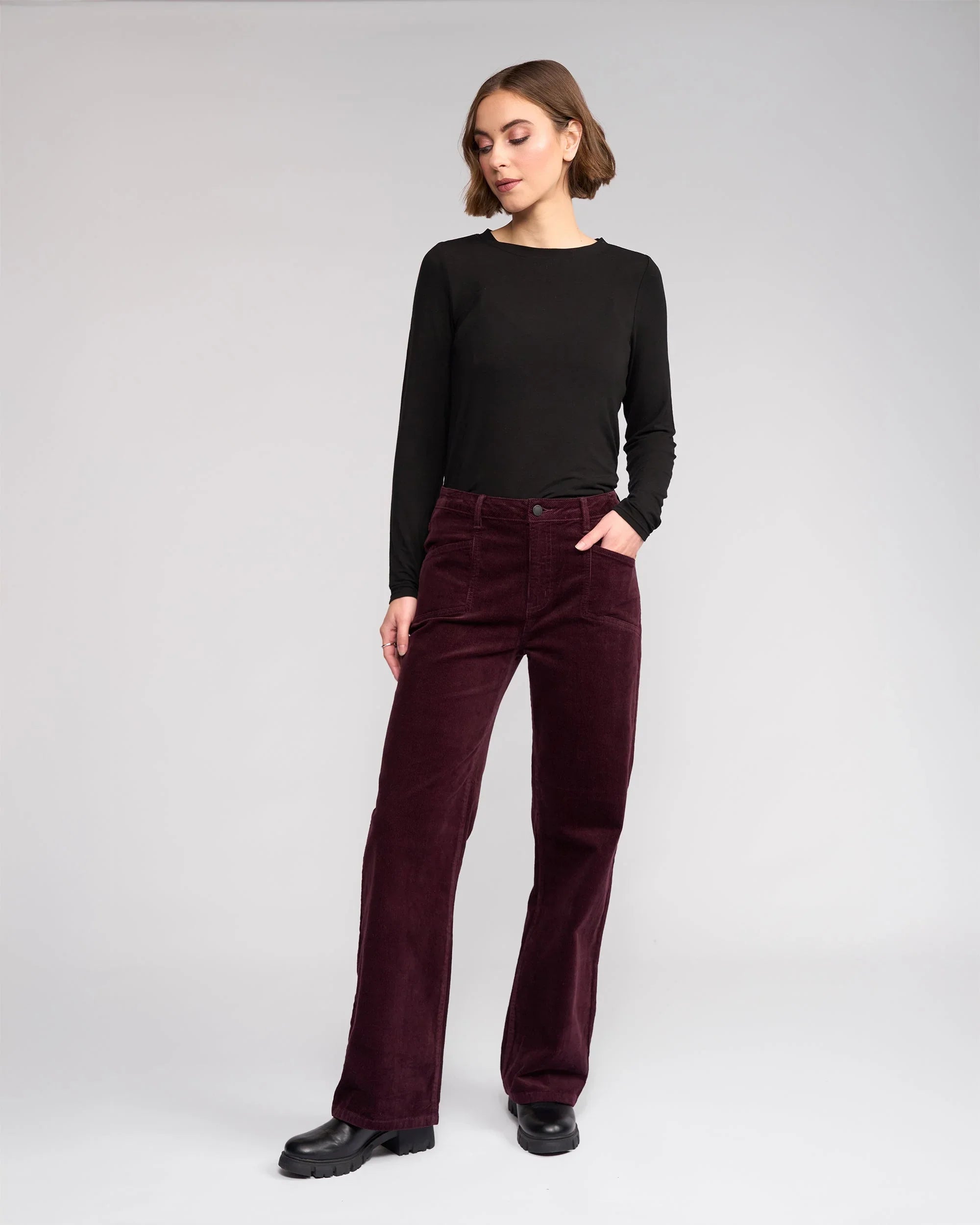 A woman stands in a studio wearing a black long-sleeve top and burgundy corduroy pants with black shoes, one hand in her pocket, looking slightly to the side against a plain background.