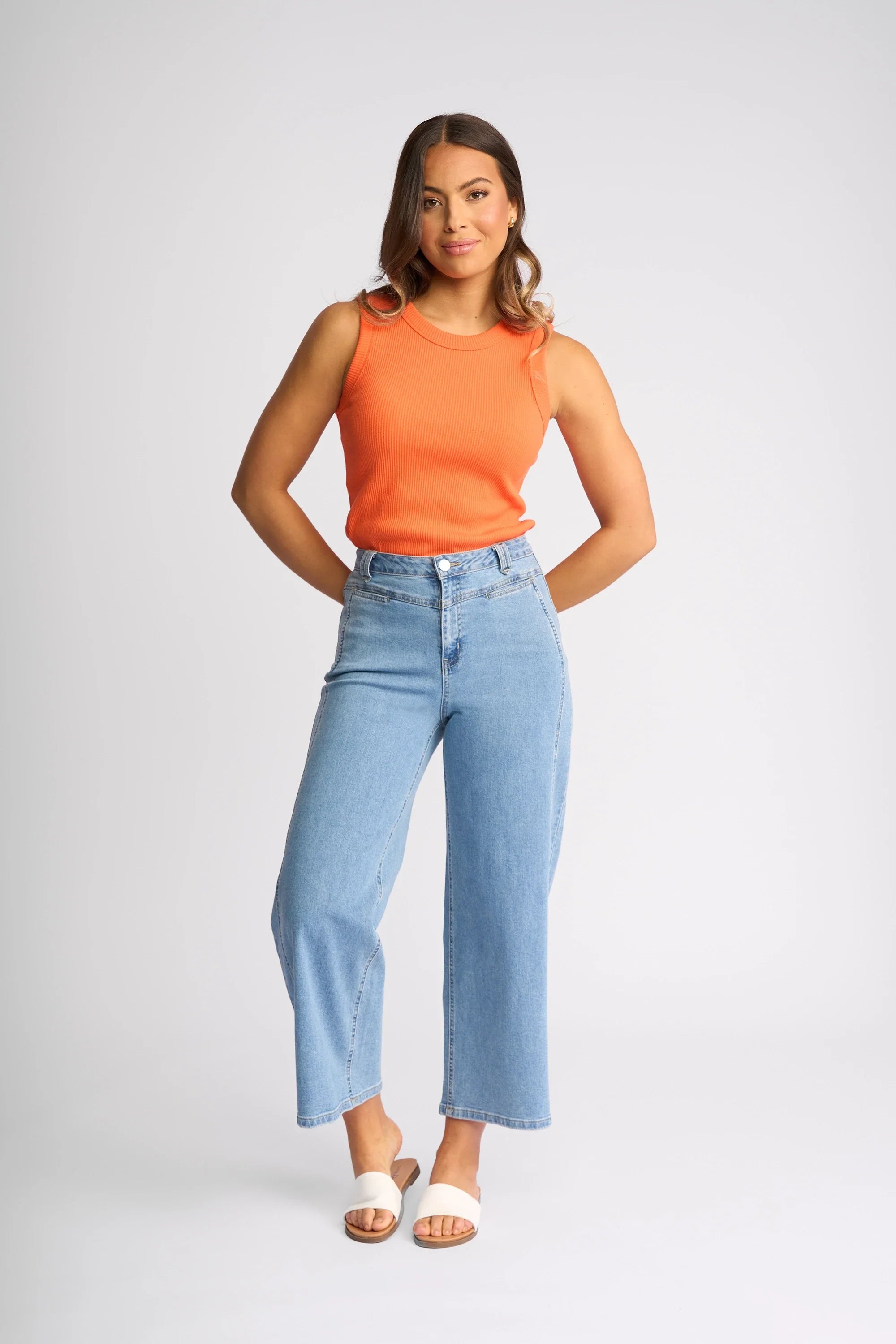 A woman stands sideways, wearing a geometric-patterned long-sleeve top, blue jeans, and brown platform shoes, against a plain light gray background.