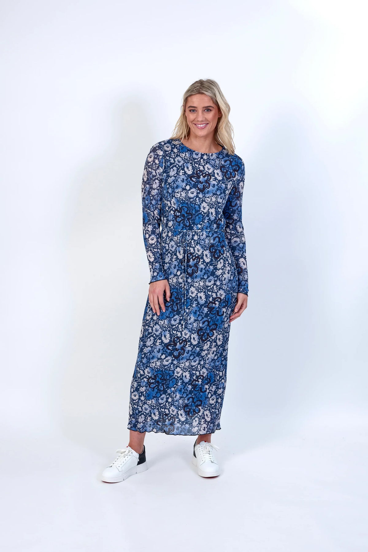 Woman wearing long sleeve blue floral print midi dress with white sneakers on white background