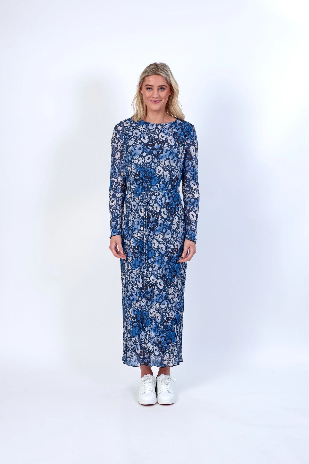 Woman in a blue floral long-sleeve midi dress with white sneakers standing against a white background