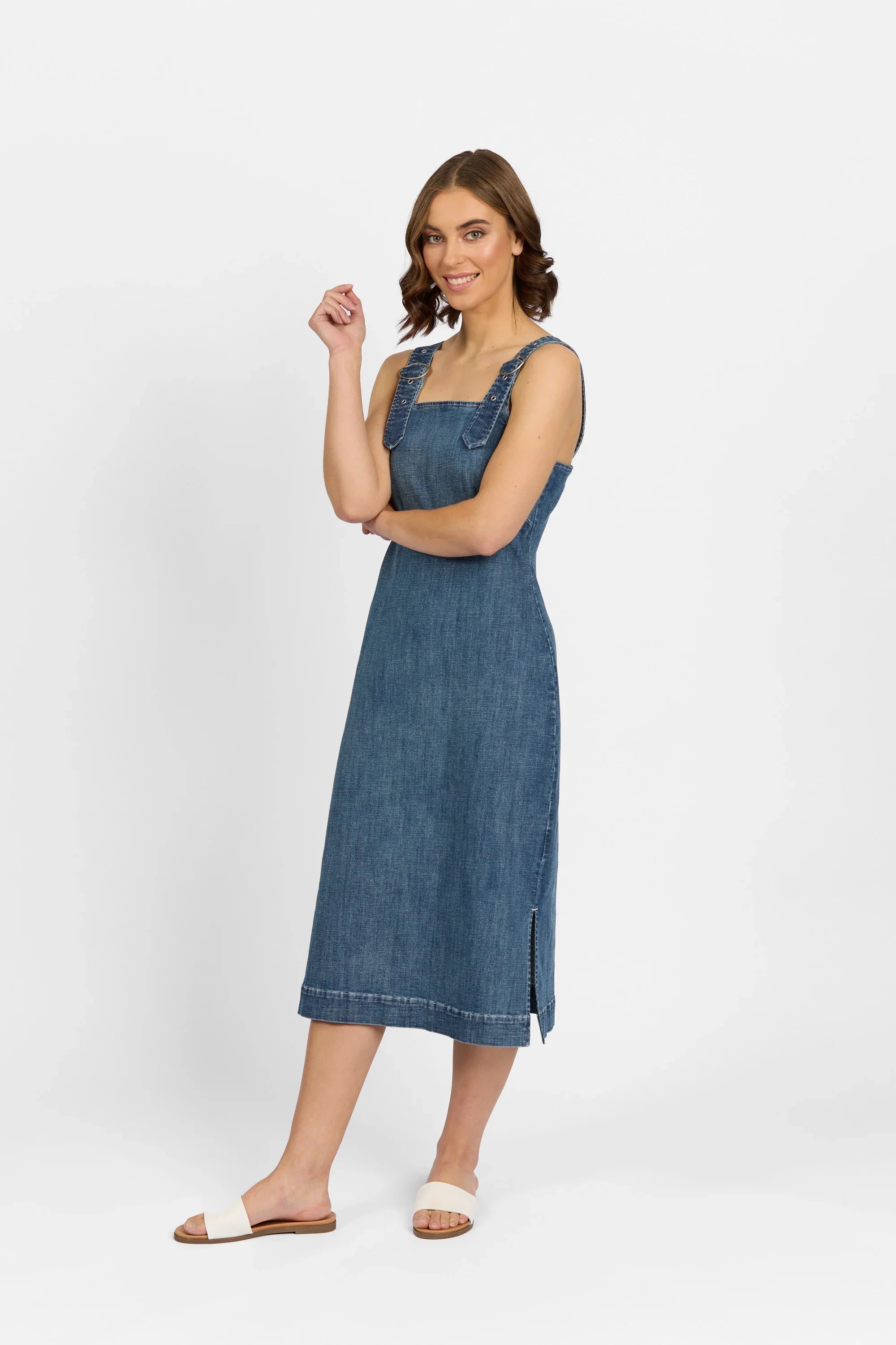 A woman stands sideways, smiling with one hand raised, wearing the Knewe Label Academy Dress - Mid Wash—a sleeveless stretch denim midi featuring side slits—and white sandals, set against a plain white background.