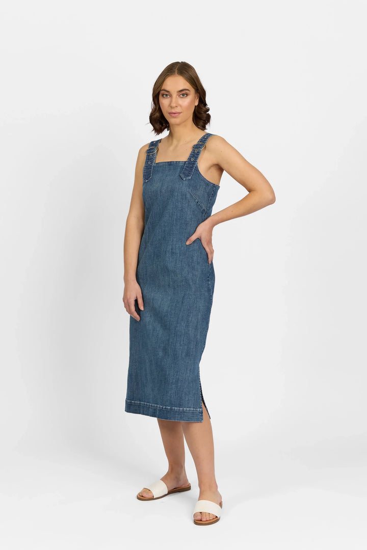 A woman with short brown hair wears the Knewe Label Academy Dress - Mid Wash, a sleeveless, knee-length stretch denim dress with wide pinafore straps, and pairs it with white slide sandals against a white background.