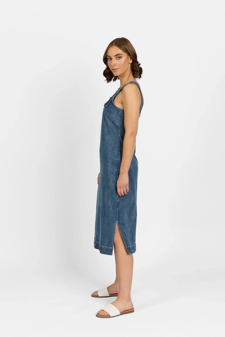 A woman stands sideways against a plain white background, wearing Knewe Label’s Academy Dress - Mid Wash, a sleeveless knee-length stretch denim dress with pinafore straps and a side slit, paired with white slide sandals.