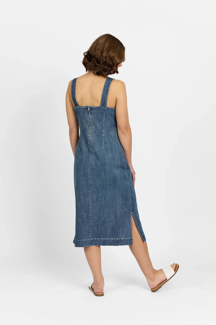 A woman with short brown hair wears the Knewe Label Academy Dress - Mid Wash, a sleeveless, knee-length stretch denim dress with a back zipper and side slit. She pairs it with white sandals and stands against a plain white background.