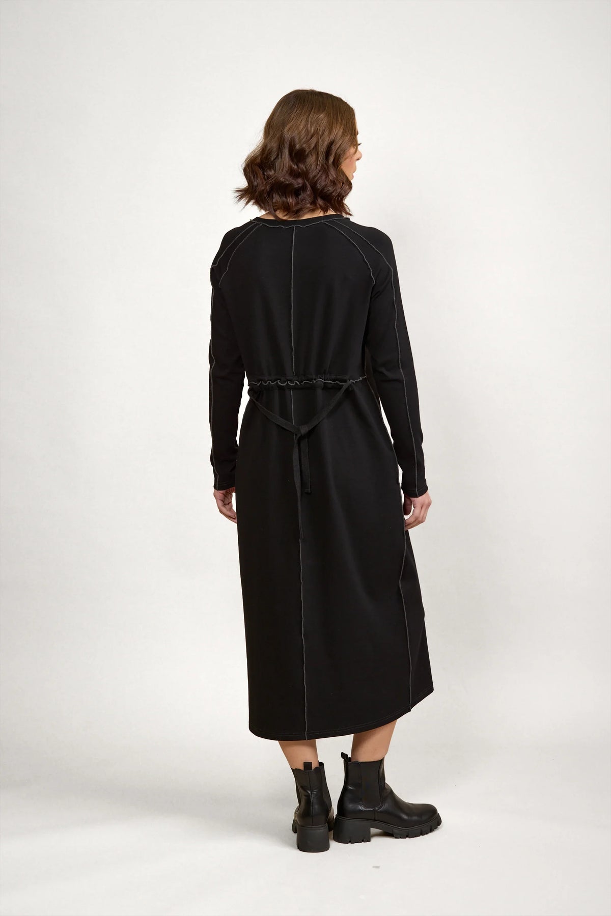 Woman in long sleeve black midi dress with visible seam details, back tie, and ankle boots