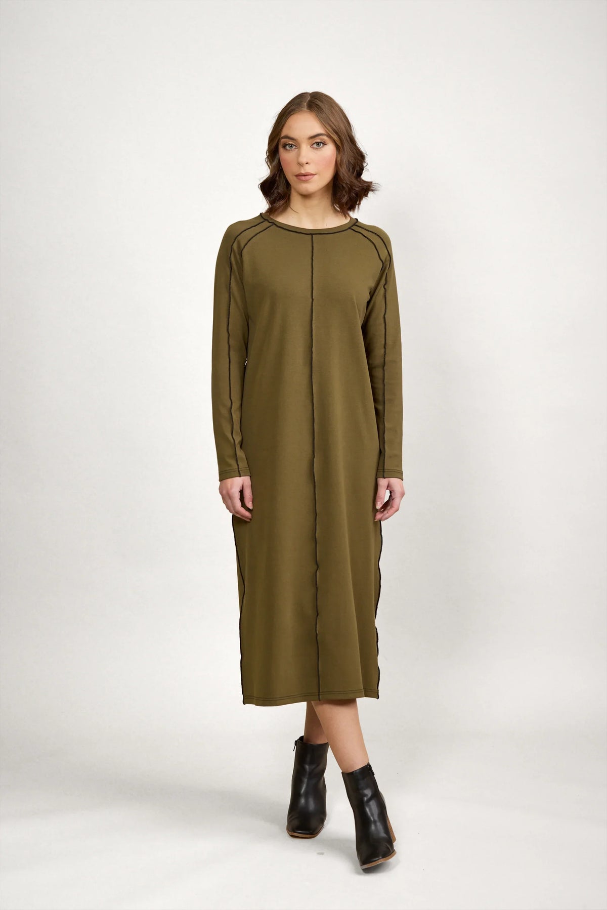 Woman wearing olive green long sleeve midi dress with black seam details and black ankle boots, standing against a plain white background.