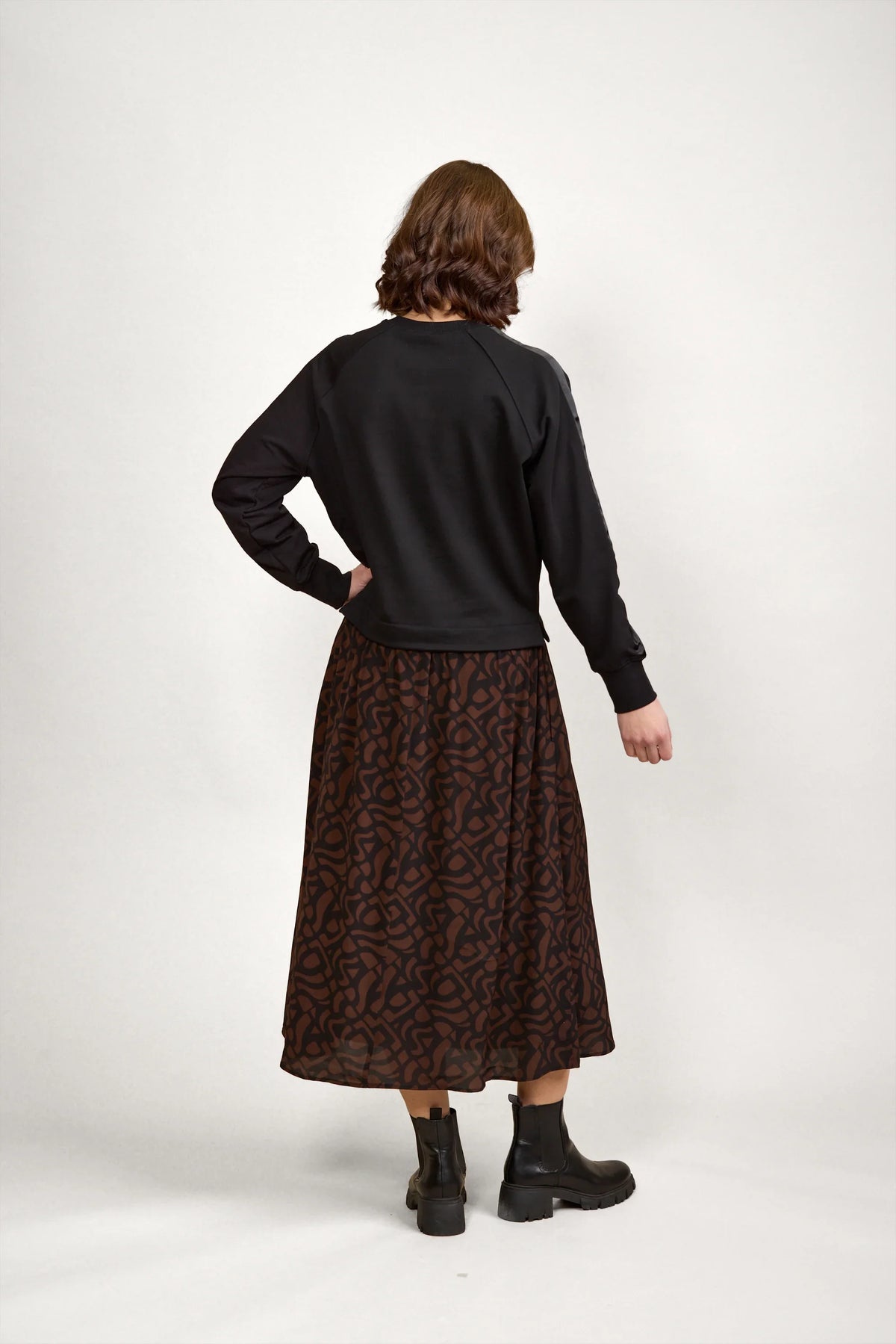 Back view of woman wearing black long sleeve top, brown abstract print midi skirt, and black boots