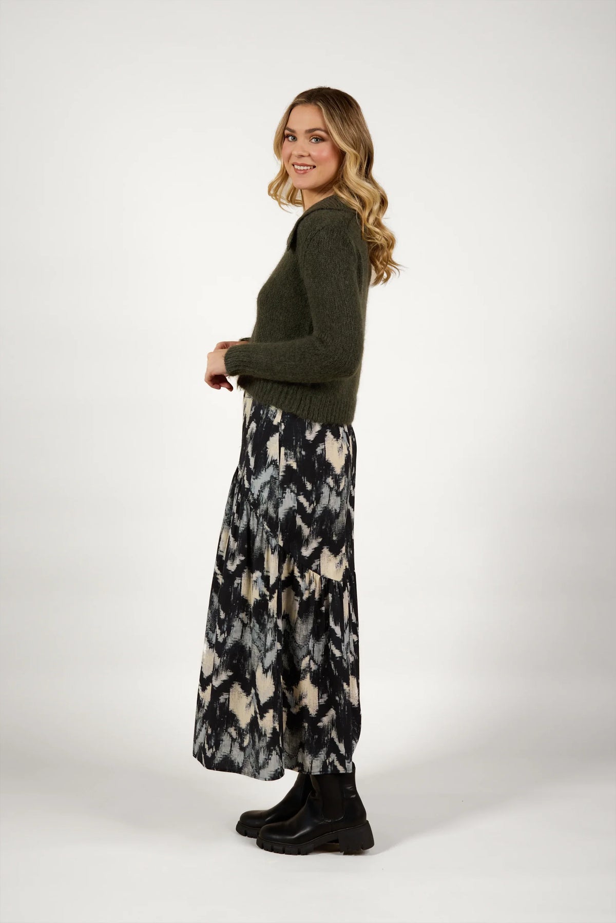 Woman in olive green knit cardigan, patterned maxi skirt, and black boots on white background