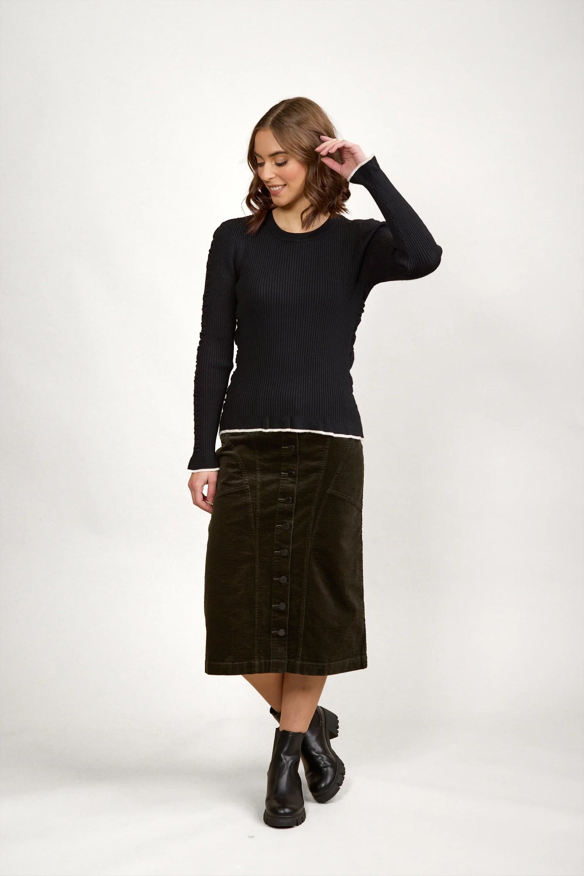 Woman wearing a black ribbed long-sleeve sweater, olive button-front midi skirt, and black boots