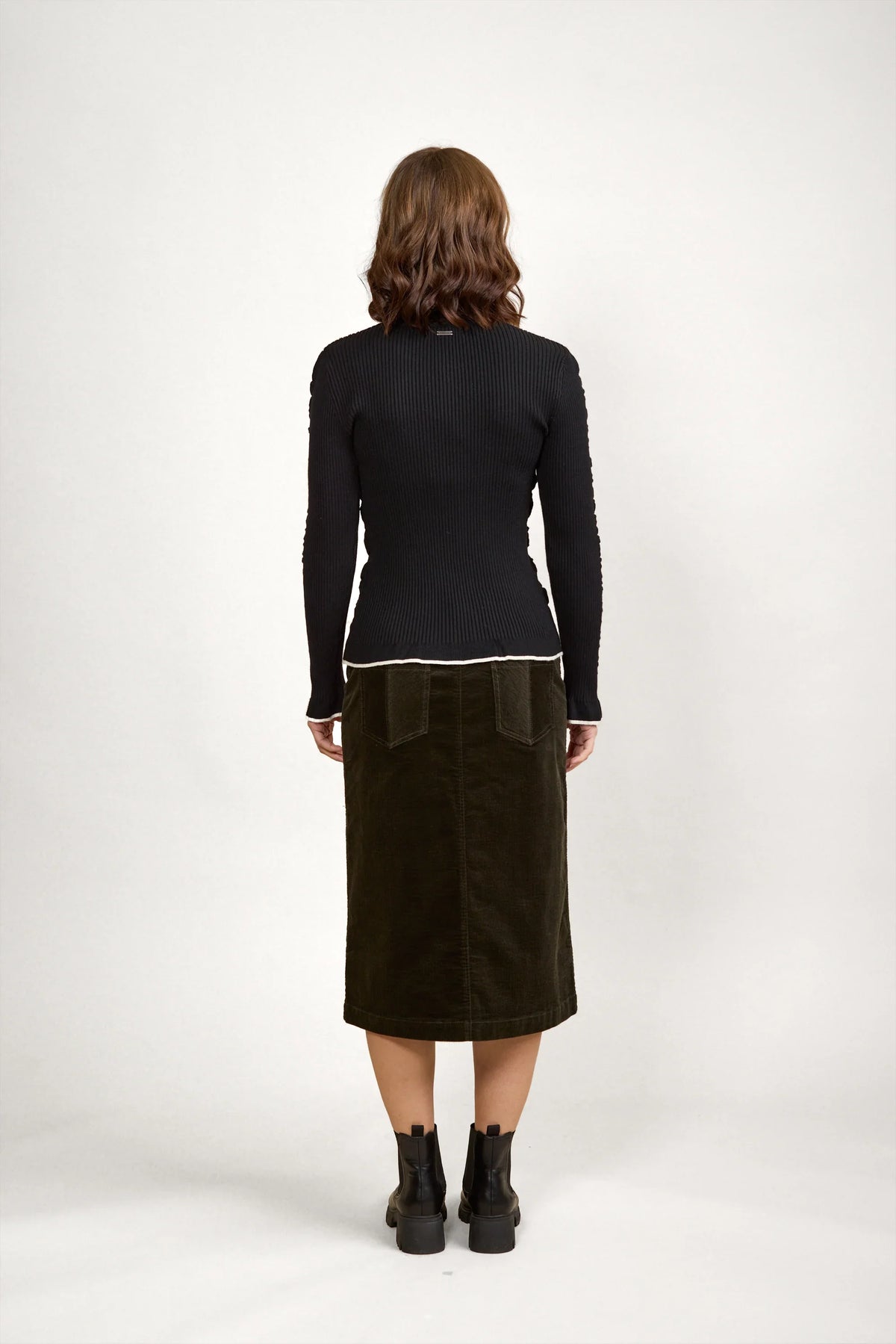 Woman facing away in black ribbed sweater, olive midi skirt, ankle boots, neutral background