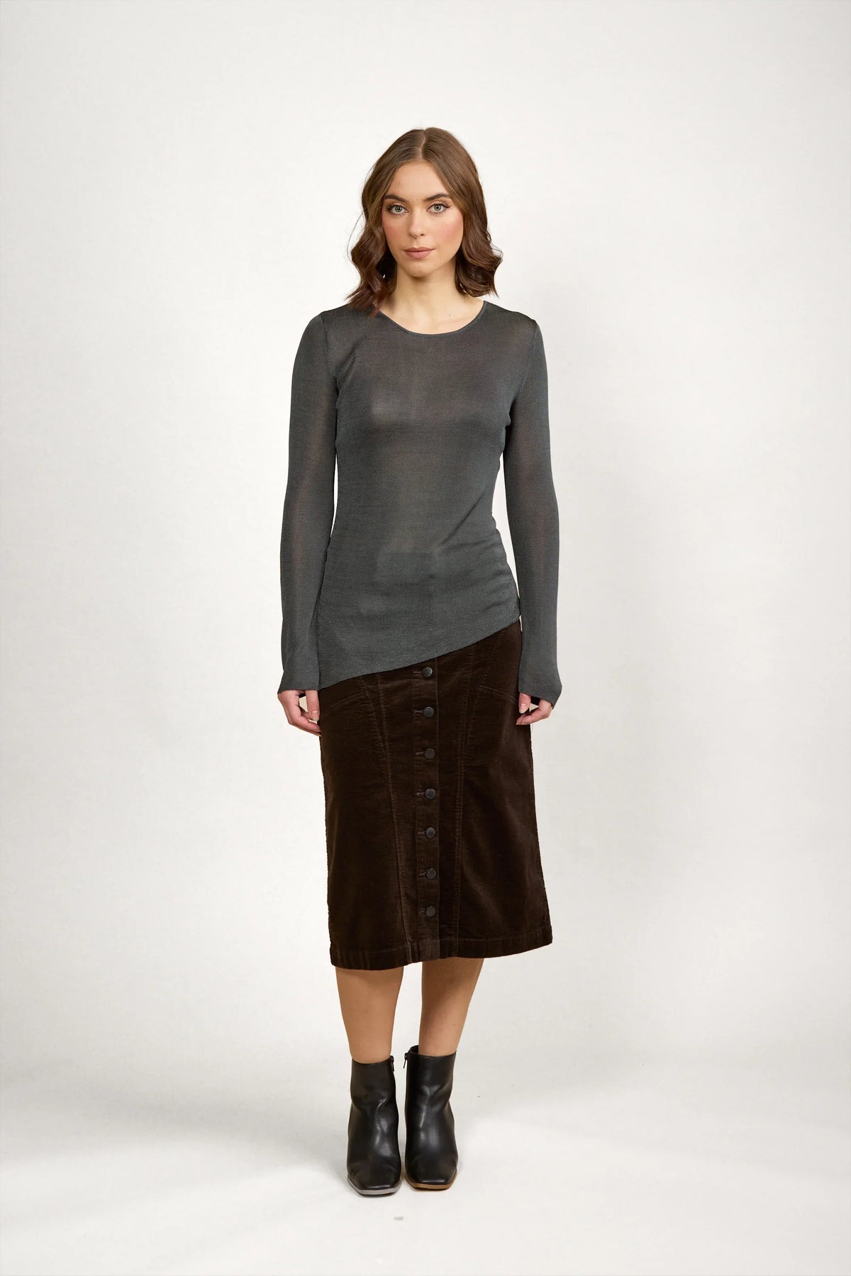 Woman modeling a sheer grey knit sweater with long sleeves, brown button-front midi skirt, and black ankle boots, standing against a white background.