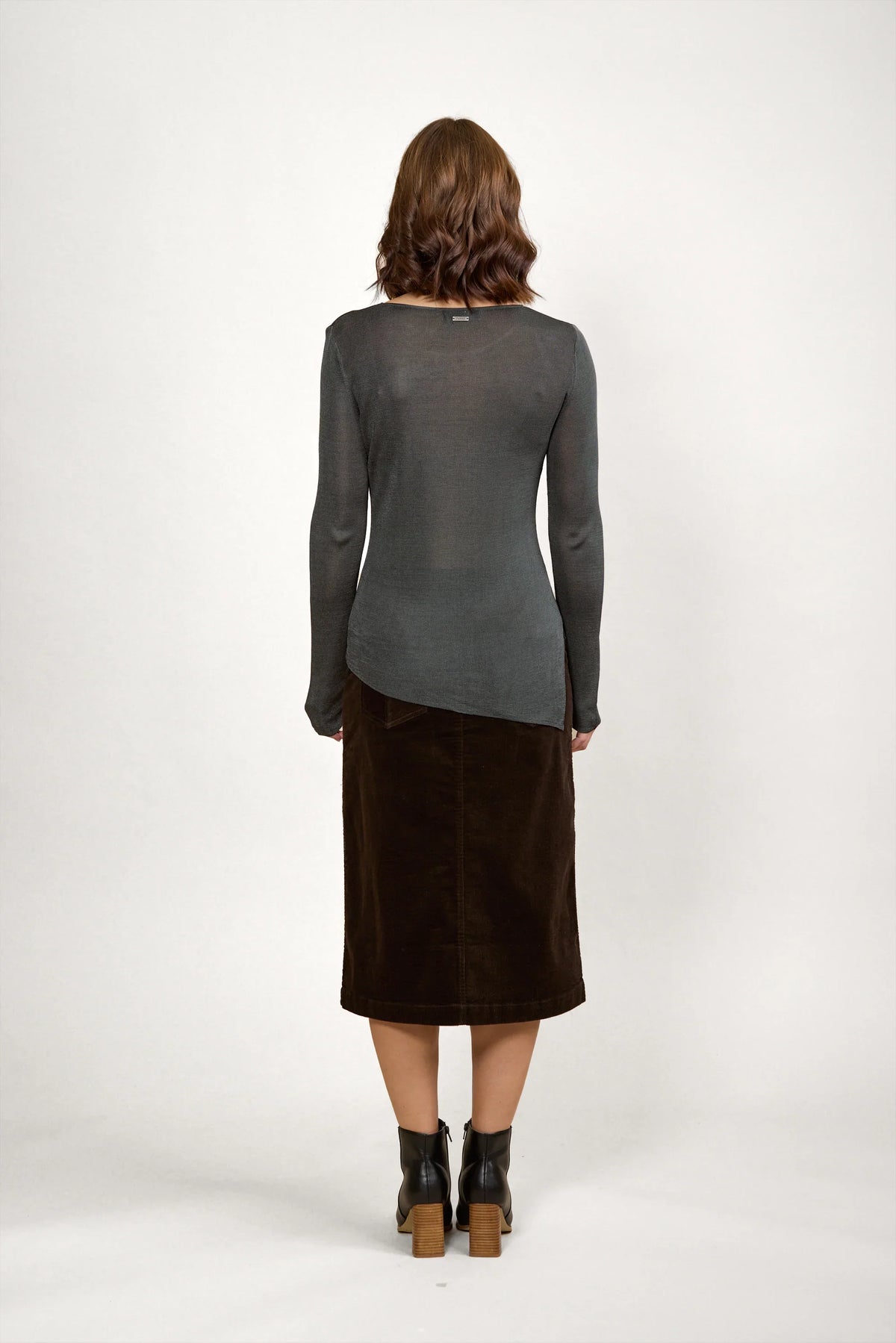 Woman in a sheer grey long sleeve sweater, brown midi skirt, and black ankle boots, back view
