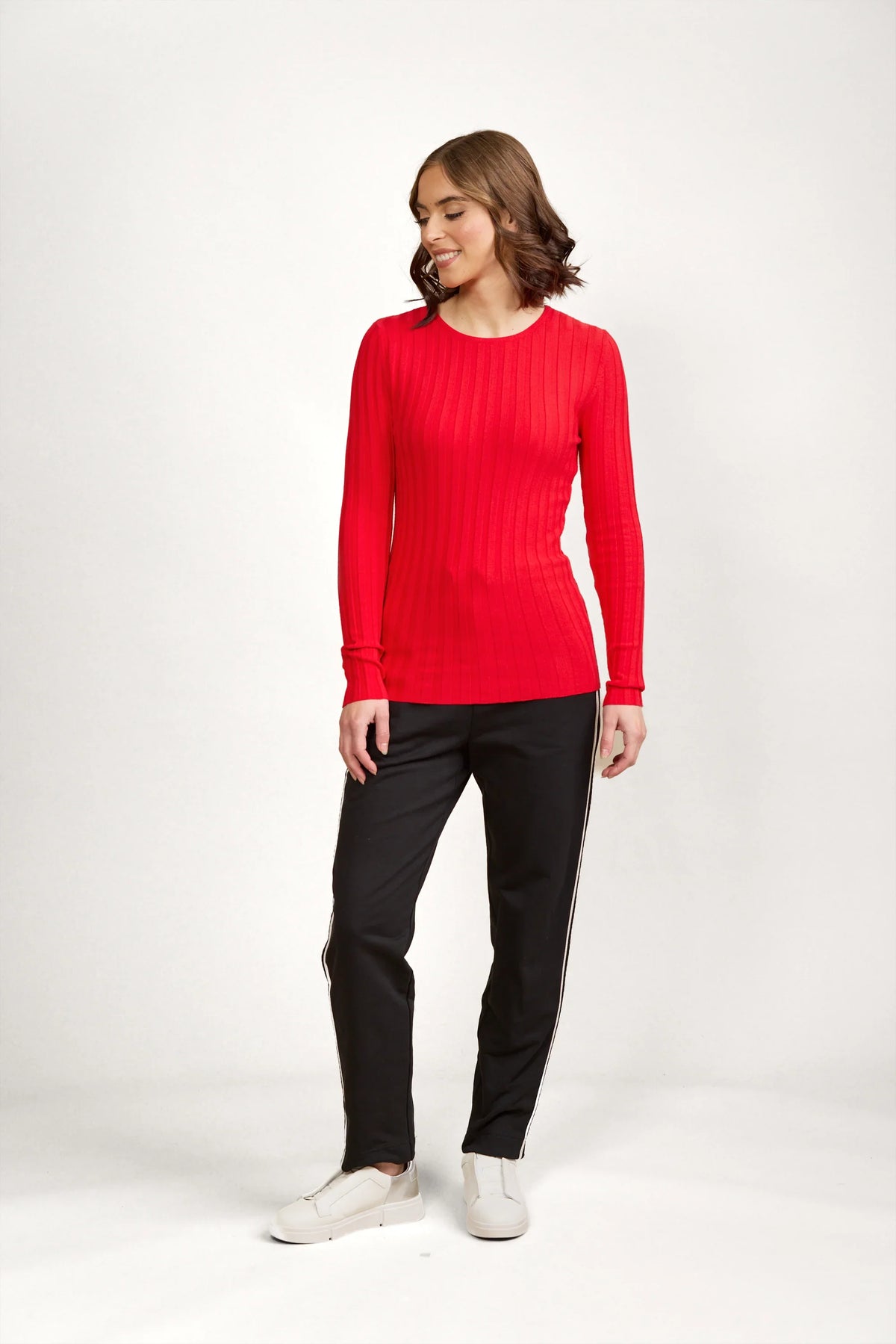Woman in a fitted red ribbed sweater with black pants and white sneakers, studio background