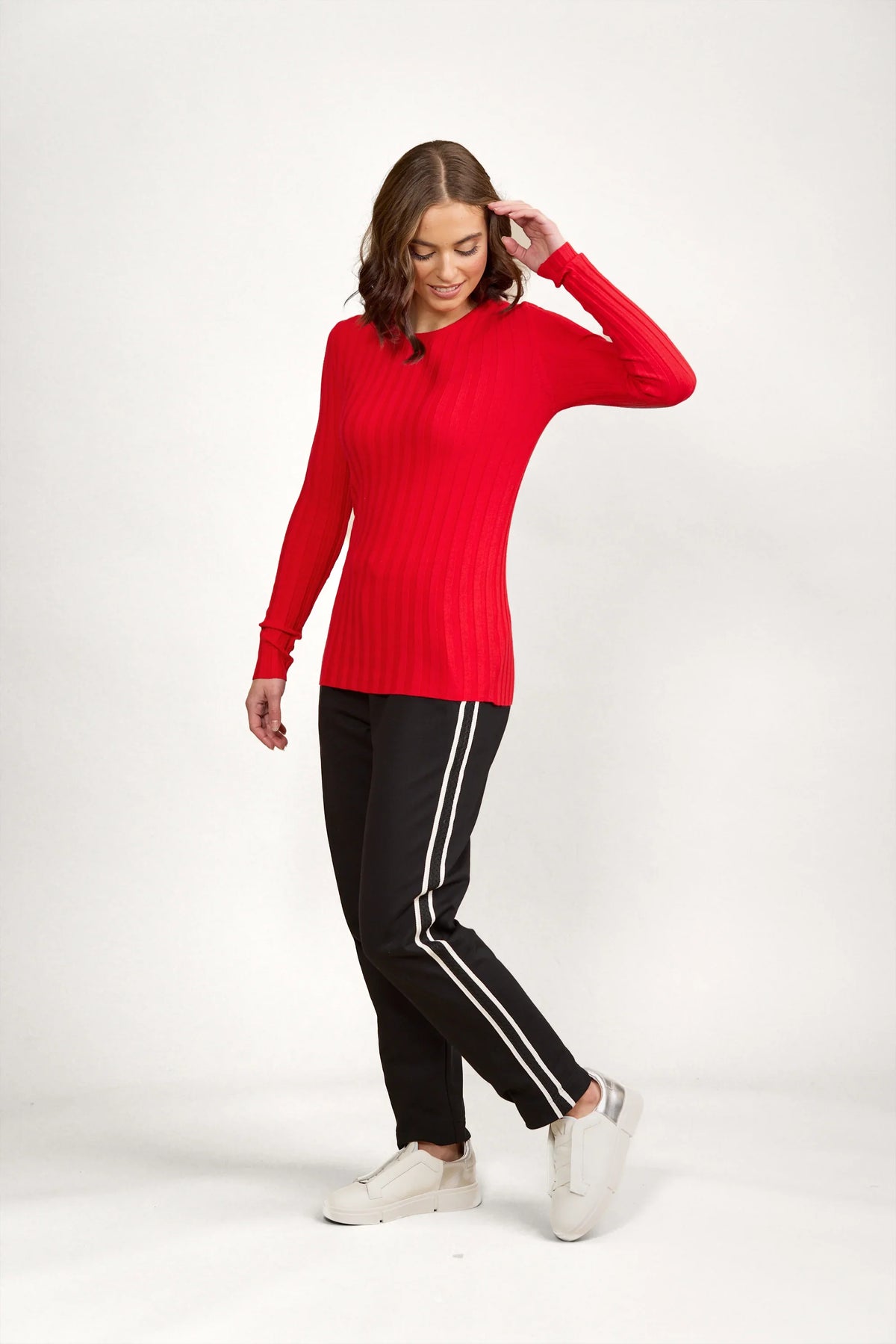 Woman in red ribbed sweater, black pants with white stripes, and white sneakers posing