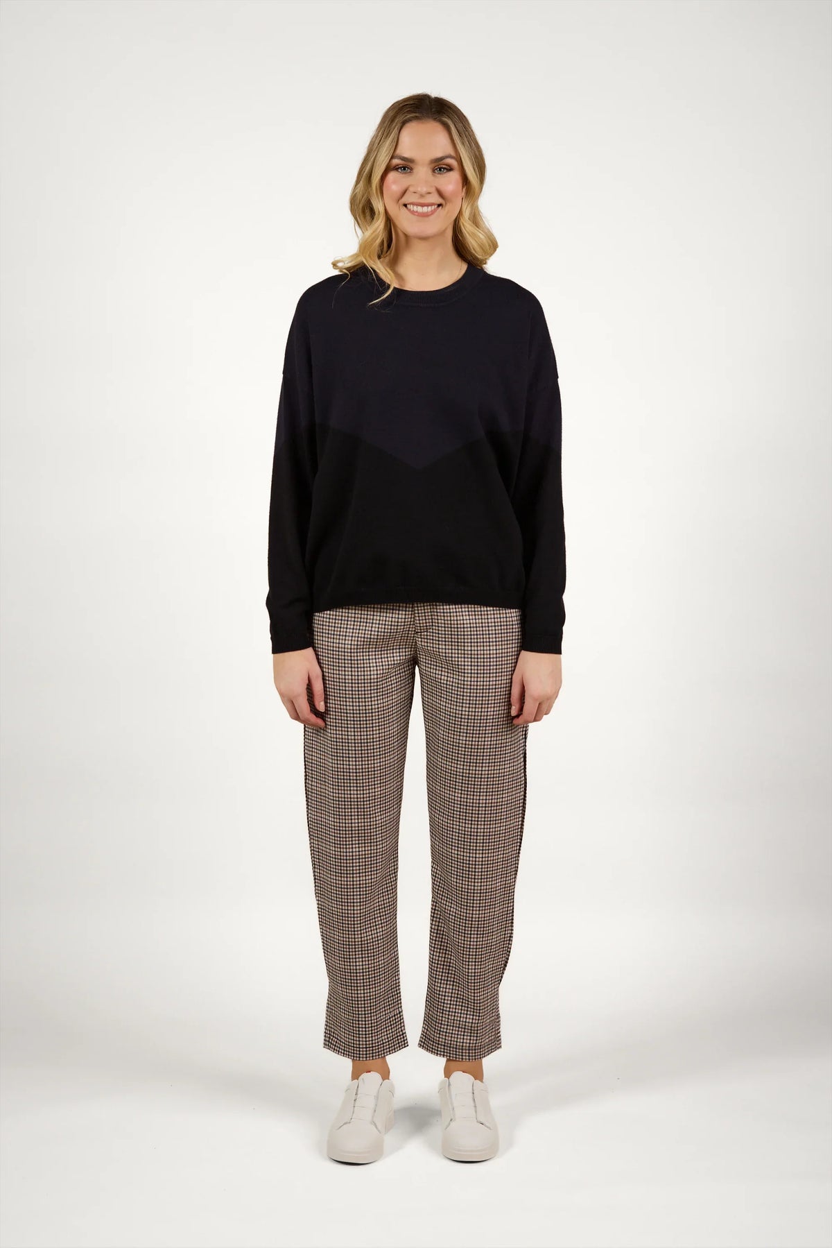 Woman wearing a black knit sweater with checkered pants and white sneakers standing against a plain background.