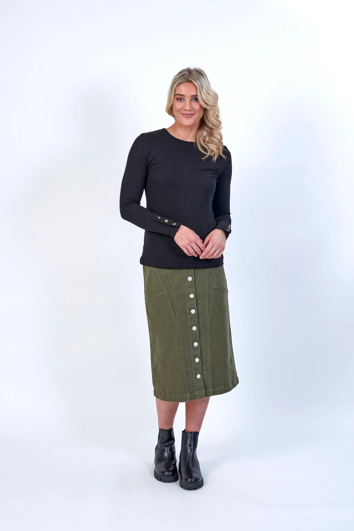 Woman wearing a black ribbed long sleeve top with gold cuff buttons and an olive green button-front midi skirt.